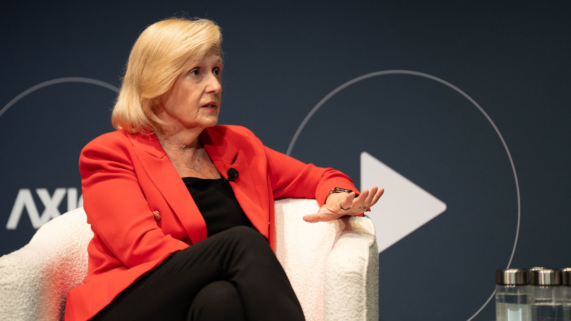 PBS CEO Paula Kerger, in a red blazer and black outfit sits on a white chair, gesturing with her hand, against a dark background with a large white play button symbol.