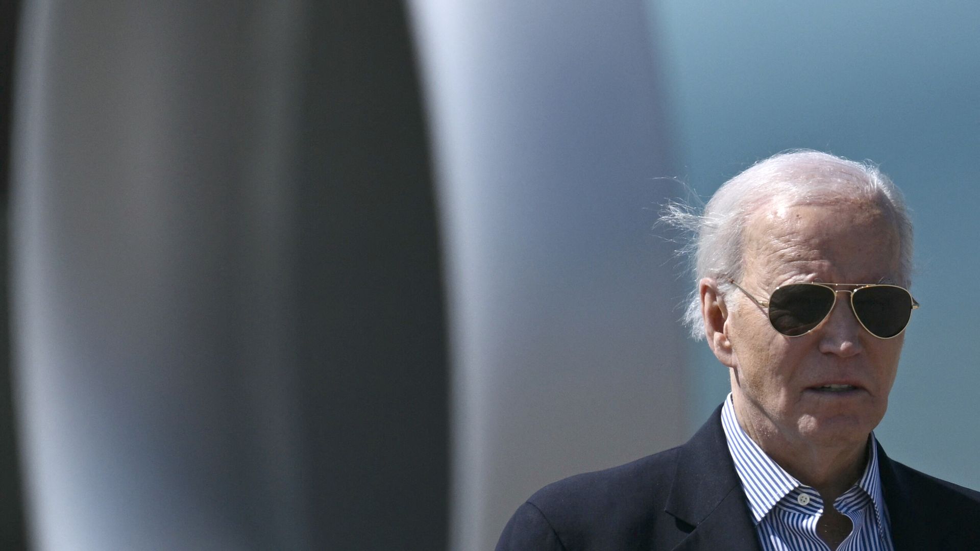 President Joe Biden, wearing aviator sunglasses, a gray suit and a white and blue shirt, stands in front of the Air Force One turbine.