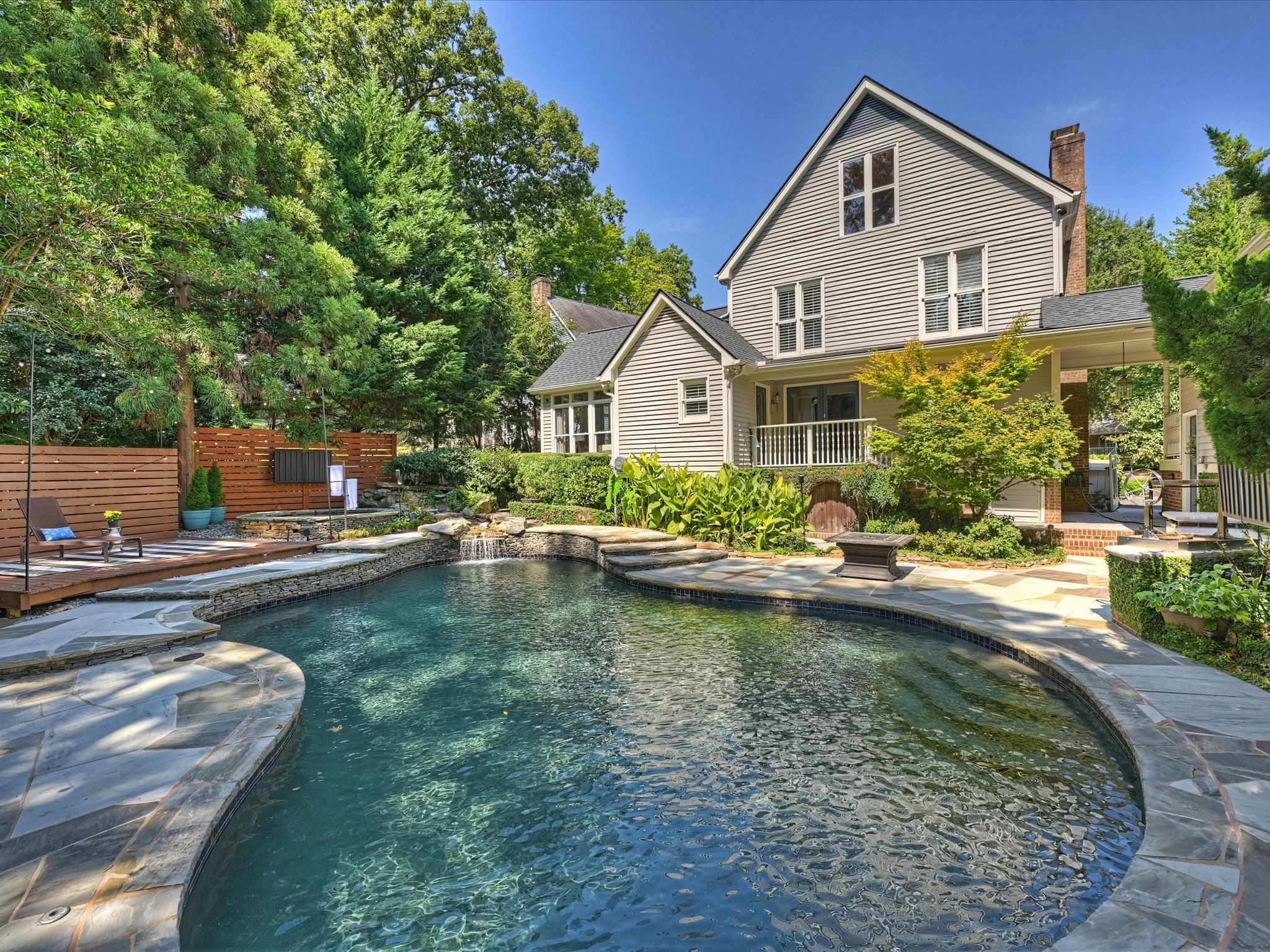 Backyard swimming pool with clear water and stone deck, surrounded by green trees and plants, adjacent to a beige two-story house with white trim and porch.