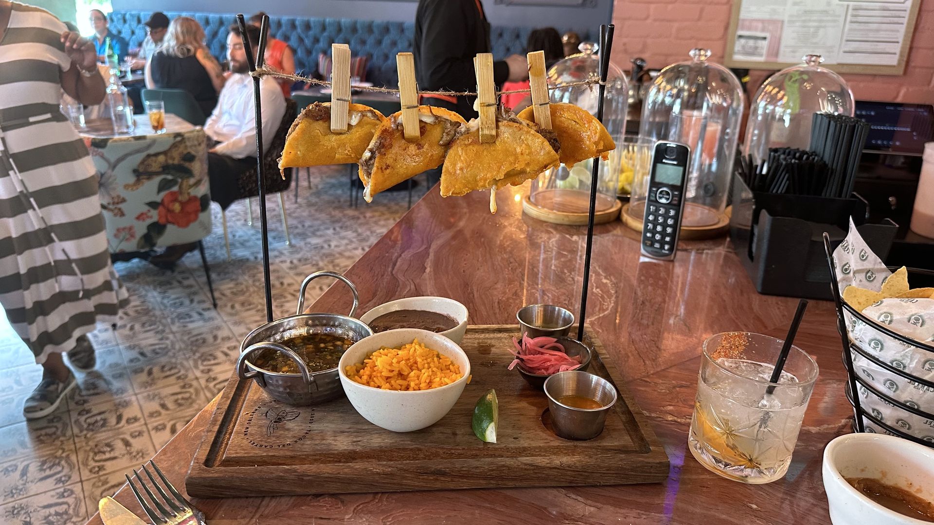 A small clotheslines has four tacos hanging from it on the corner of a bar top.