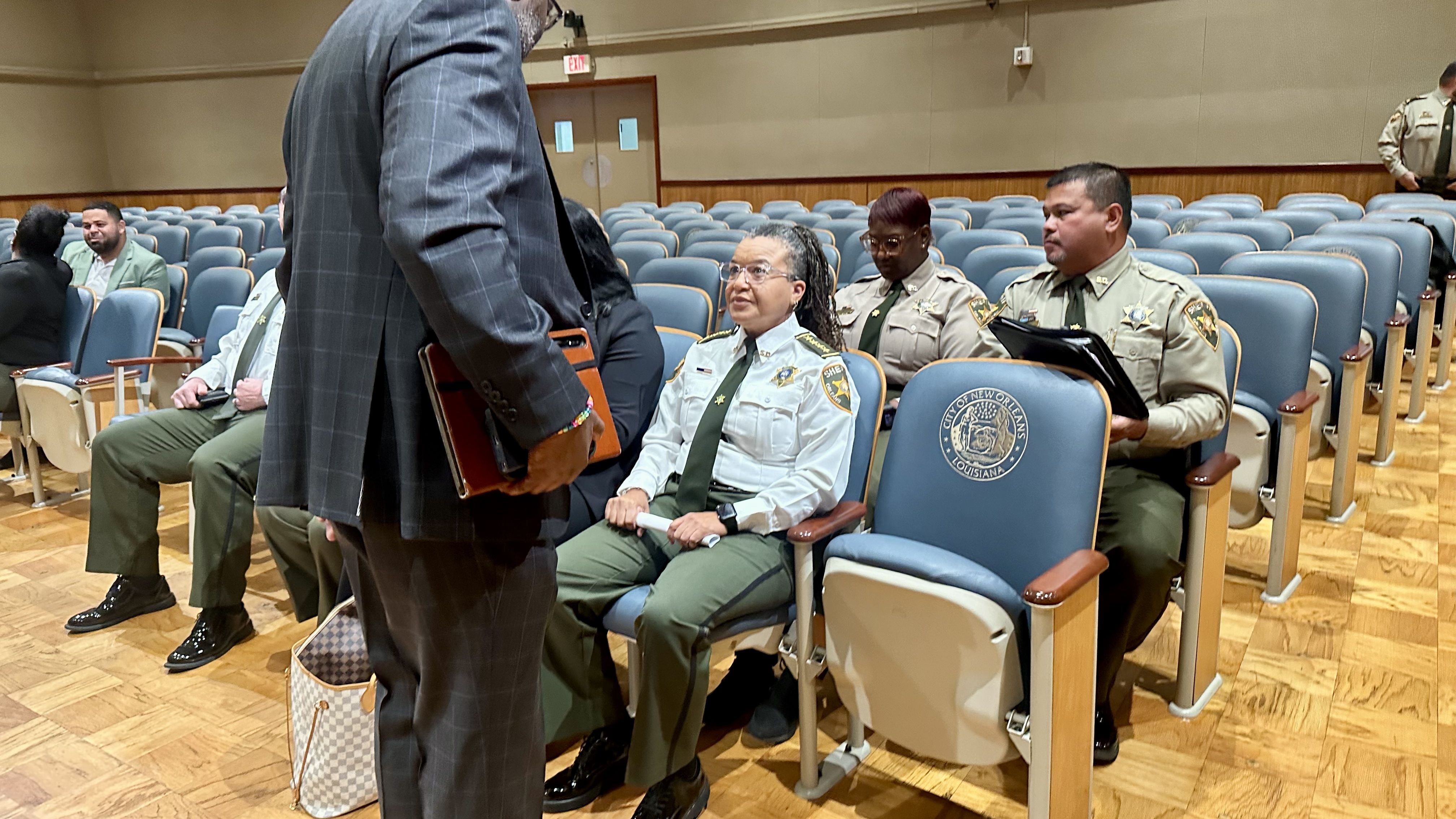 Auditorium with blue seats; uniformed officers sit in rows, including a braided woman in a light blue shirt and green tie. A suited man in foreground holds a folder; chair bears New Orleans emblem.