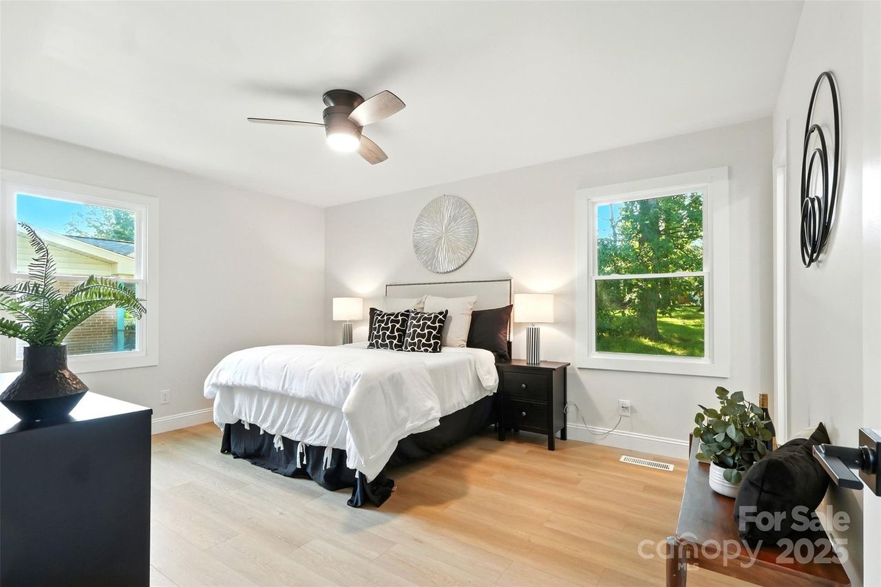 Bright bedroom with white walls, light wood floor, white bed with black and white pillows, two black nightstands with lamps, ceiling fan, and two windows showing green trees outdoors.