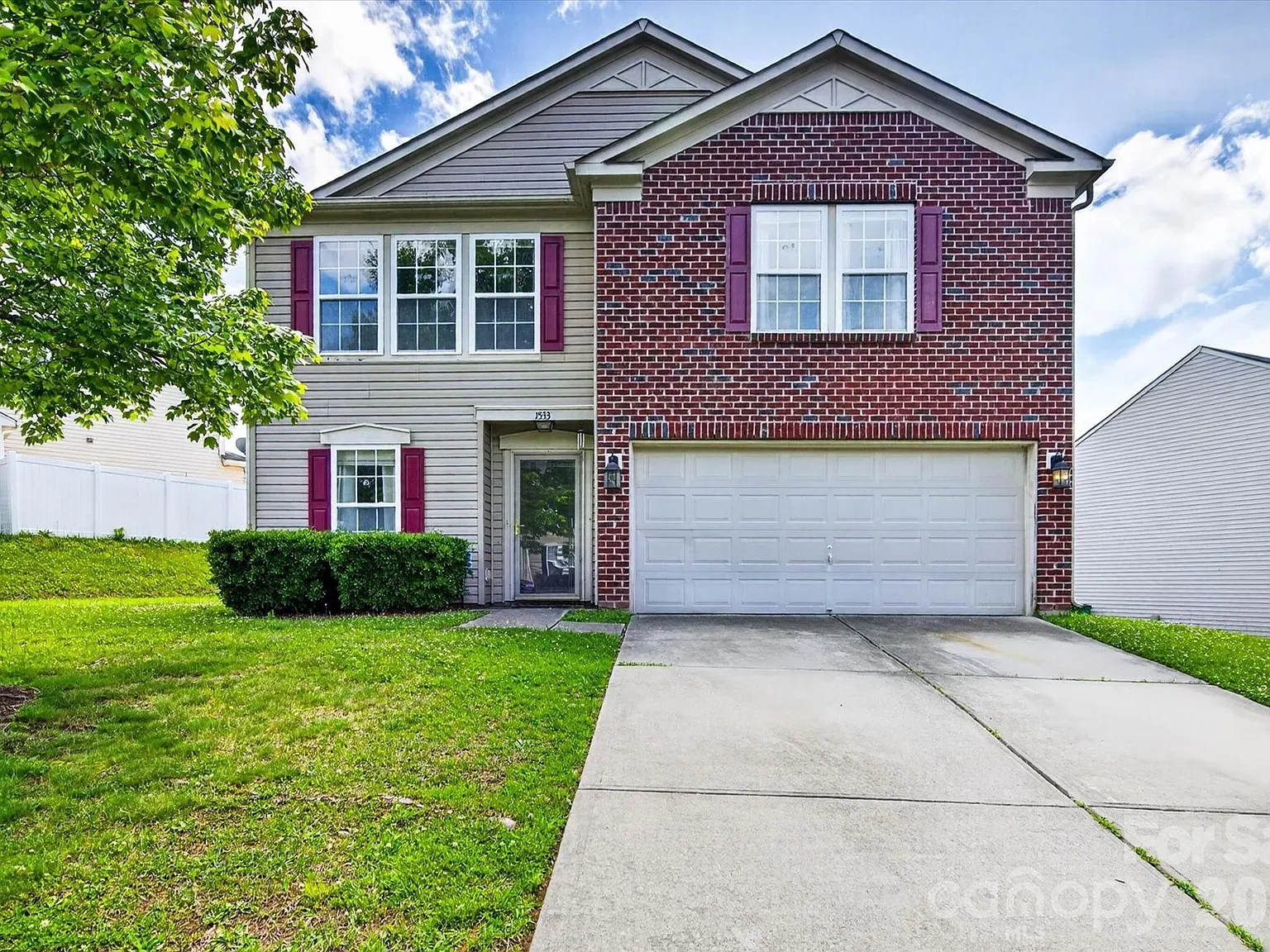 Two-story suburban house with brick and beige siding, purple shutters, and a white two-car garage. A front entry between brick and siding, large front windows, manicured bushes, green lawn, and a tree to the left under a blue sky.