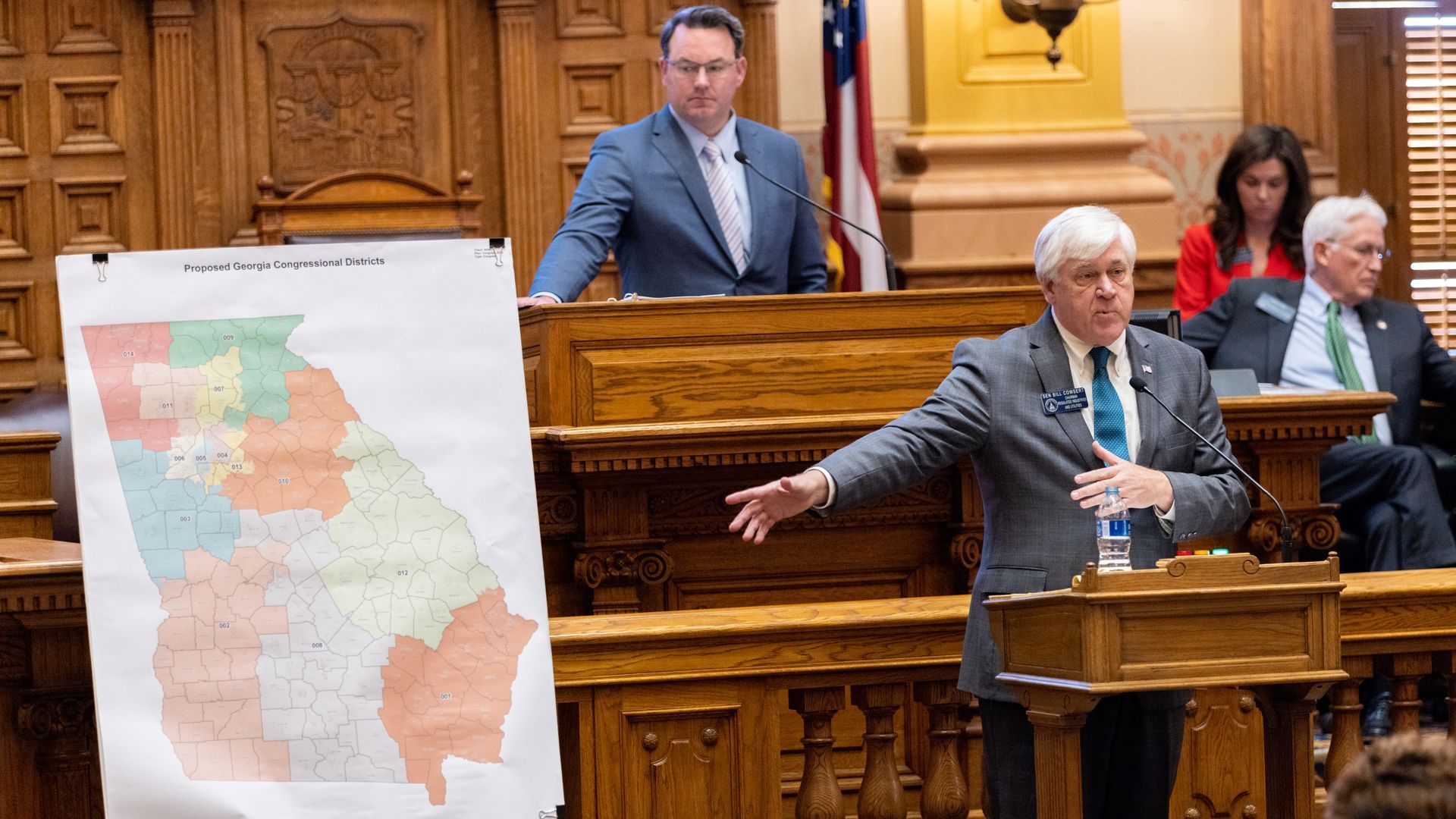 A man in a suit standing next to a large political map of Georgia speaks in the well of a legislative building
