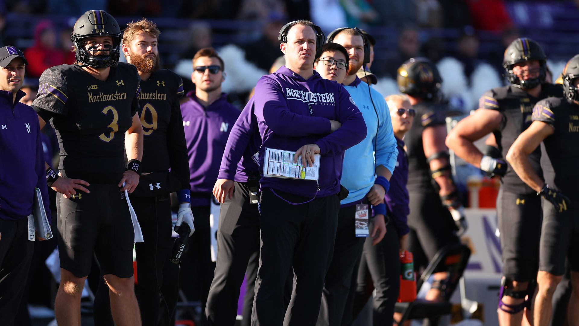 Coach in purple hoodie with arms crossed, surrounded by football players in black uniforms and helmets.
