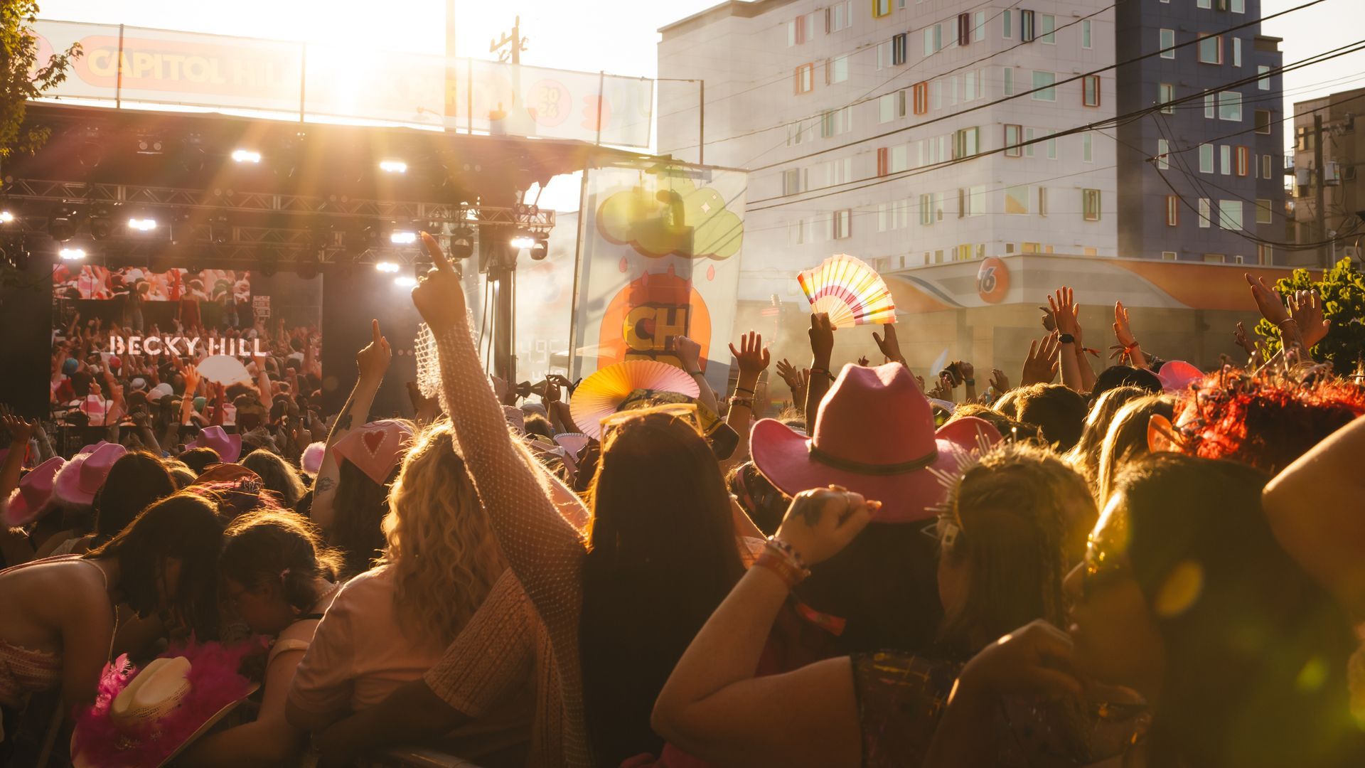 A crowd of people stands in front of a stage as the sunlight fades away, with one person at the center raising their hand up.