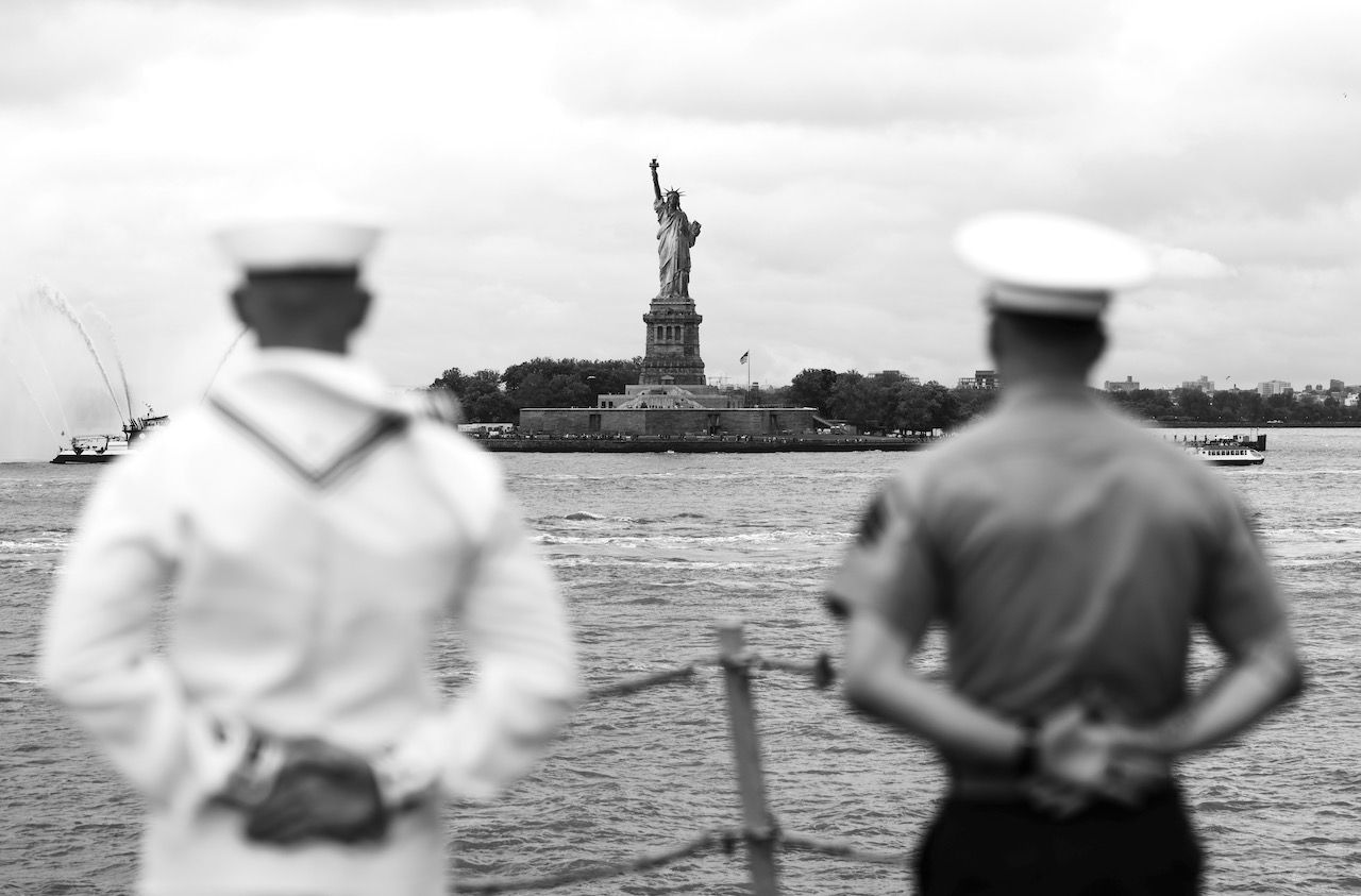 Two sailors stand with hands behind backs, blurred foreground, watching the "Statue of Liberty" across the harbor; black-and-white scene with boats and distant cityscape.