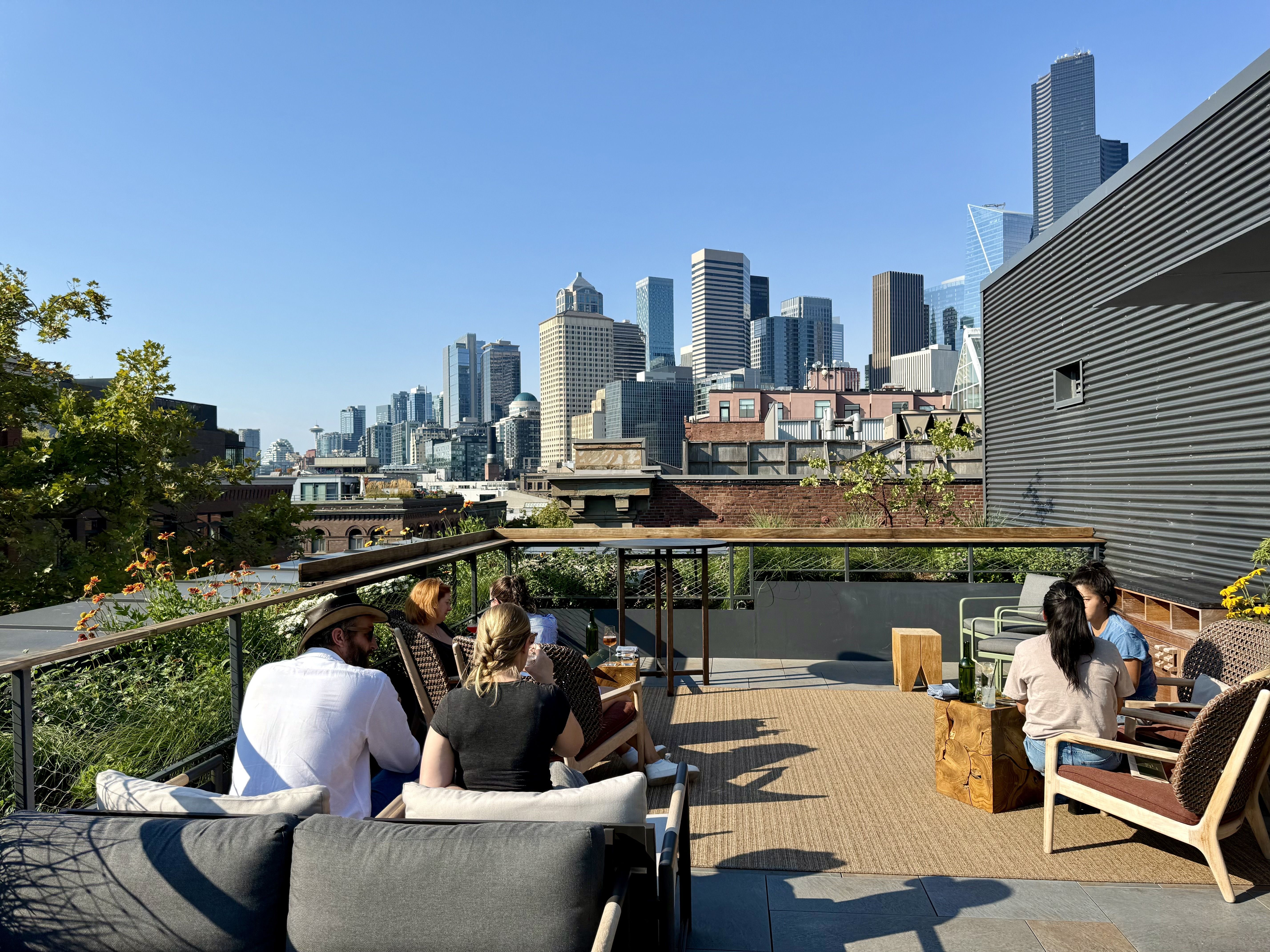People sitting and chatting on a sunny rooftop patio with wooden furniture and Seattle city skyline in the background under a clear blue sky.