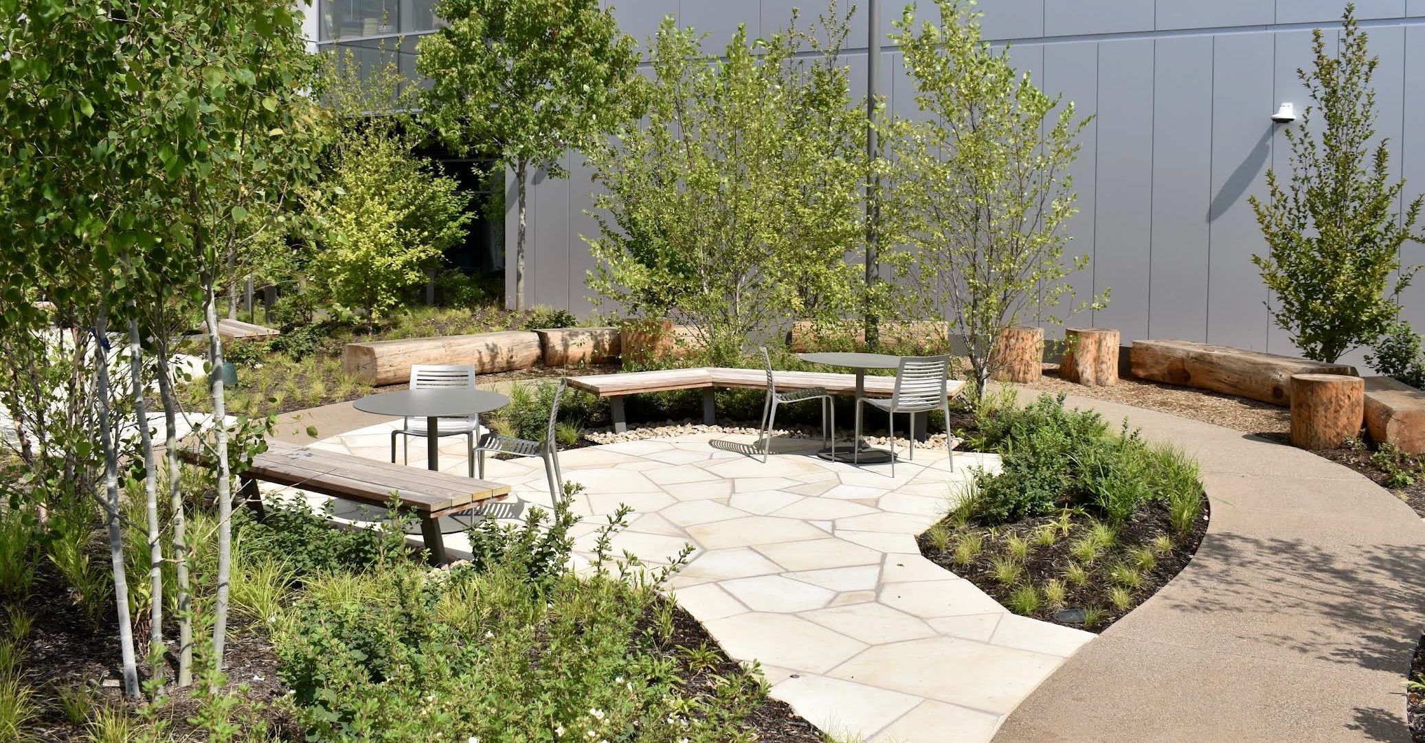 Modern outdoor courtyard with gray panel building, lush trees and shrubs, wood log benches, and round gray tables with matching chairs on pale stone pavers.