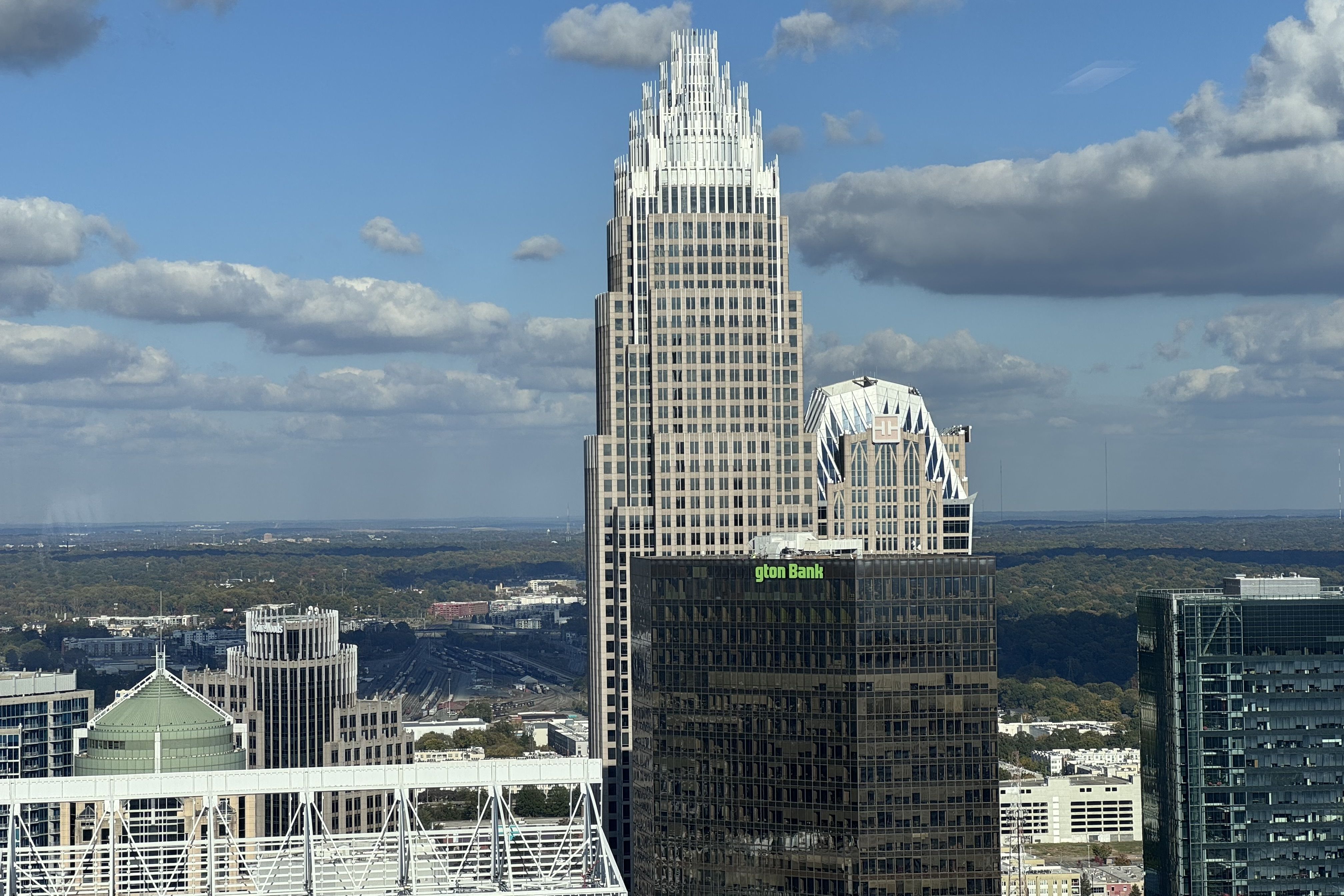 Prominent skyscraper with a stepped white crown and dark glass facade rises above surrounding buildings, set against a blue sky with scattered clouds; a green bank sign is visible at the base.