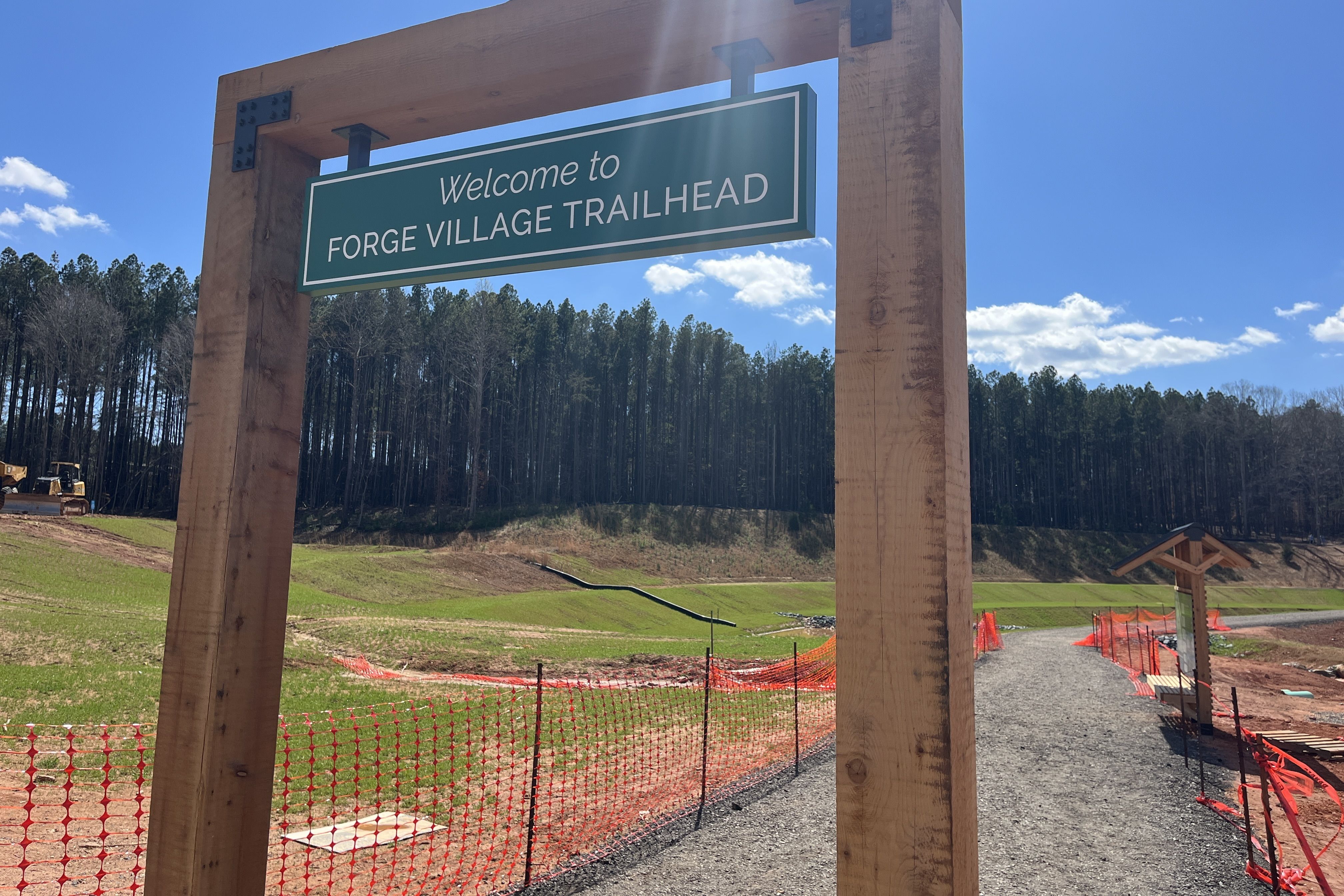 Wooden frame sign reads "Welcome to FORGE VILLAGE TRAILHEAD" over a gravel path, with pine trees in the background, a blue sky, and orange construction fencing along the trail.