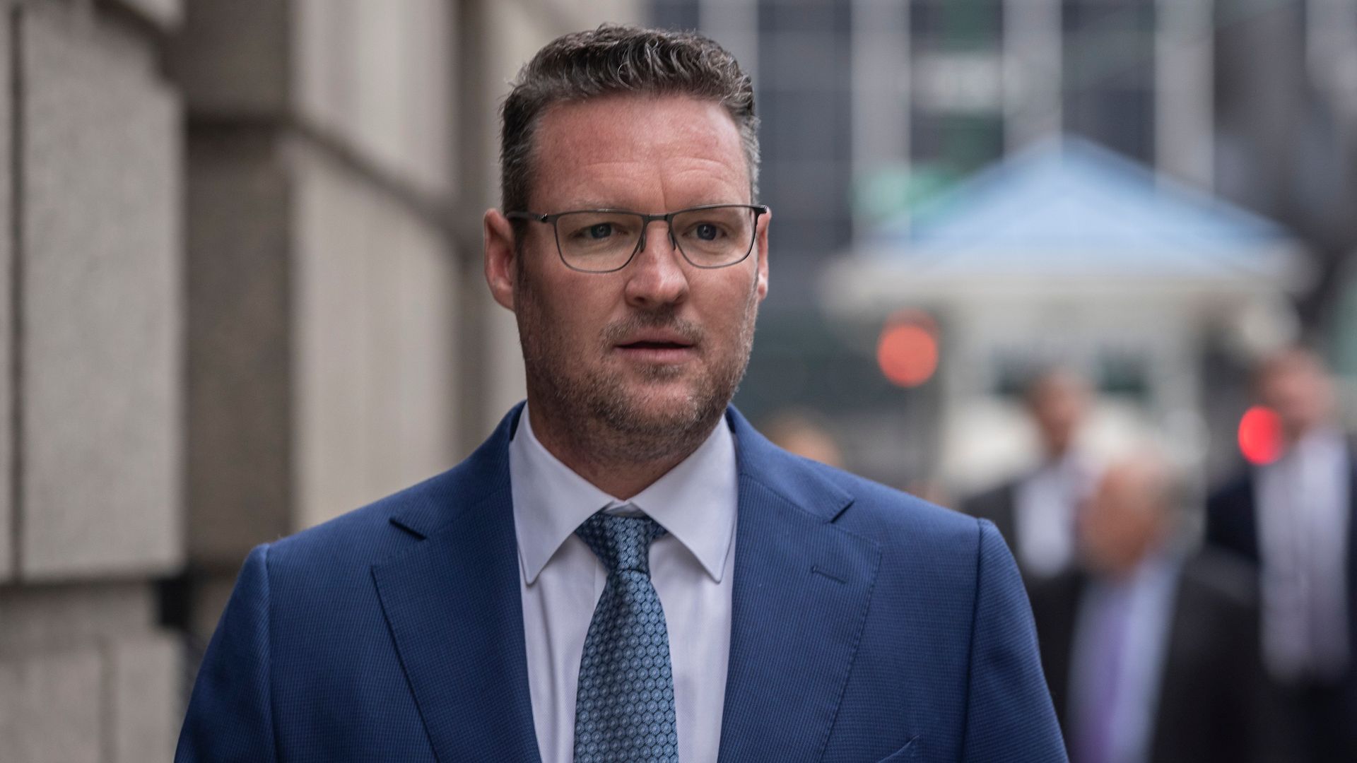 A head-and-shoulders photo of a white man with short curly brown hair and a blue suit, standing on a city street.