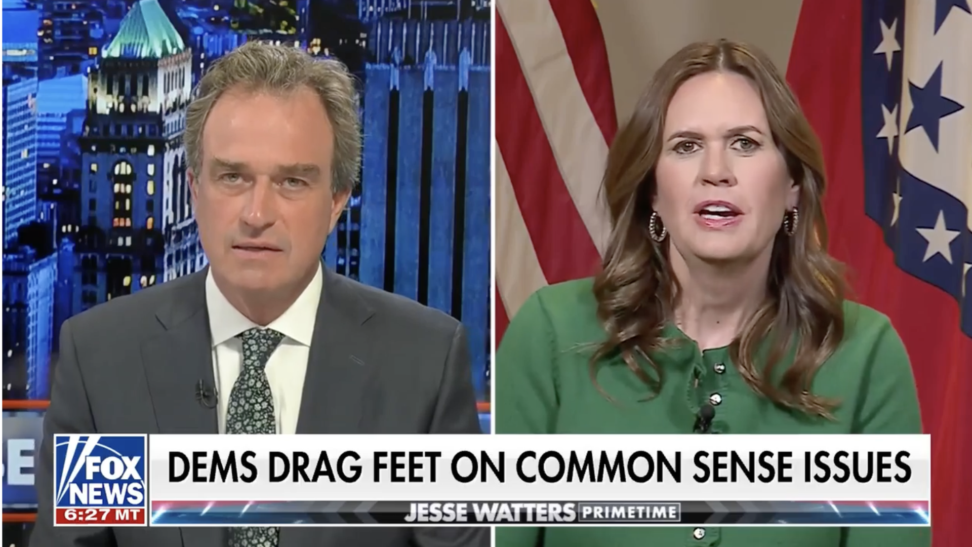 Split-screen TV interview: a male host on the left and a woman speaking on the right, with U.S. and Arkansas flags behind her.