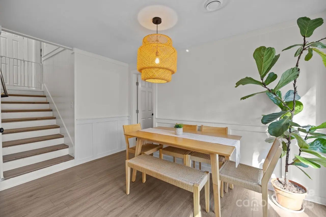 Bright dining area with light wooden table, chairs, and bench under a yellow woven pendant light; green potted plant by sunlit window and white walls with staircase nearby.