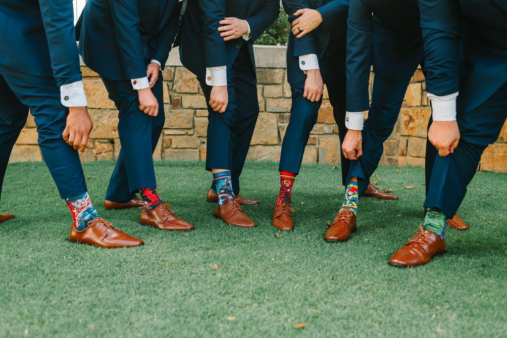 Five men in navy suits and brown shoes showing colorful patterned socks outdoors on green grass against a stone wall background, bending to reveal their socks.