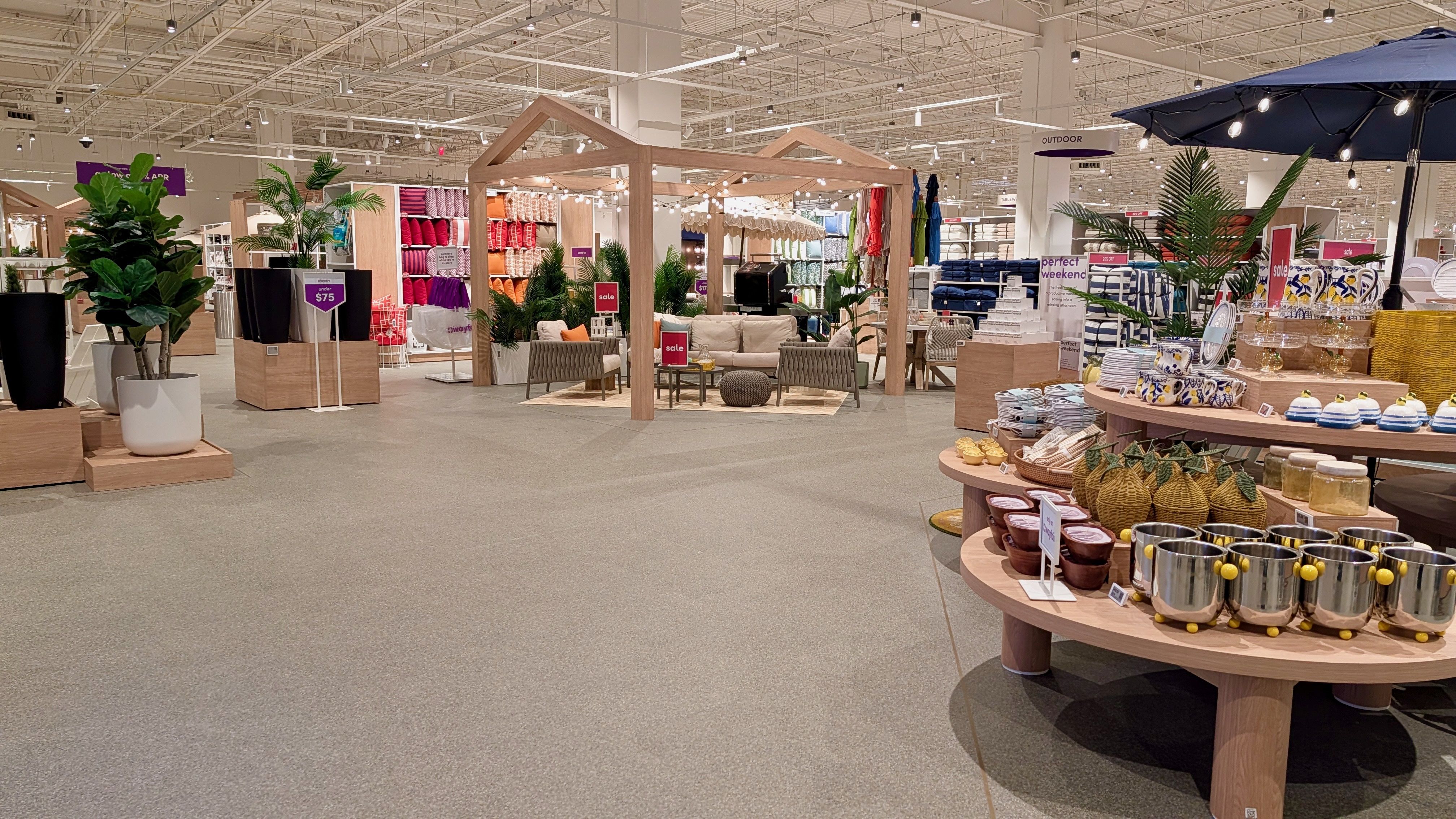 Bright home goods store with a wooden pergola display in the center, beige sofas around a coffee table, potted plants, and a round wooden table on the right with mugs and bowls; sale signs throughout, colorful textiles on back shelves.