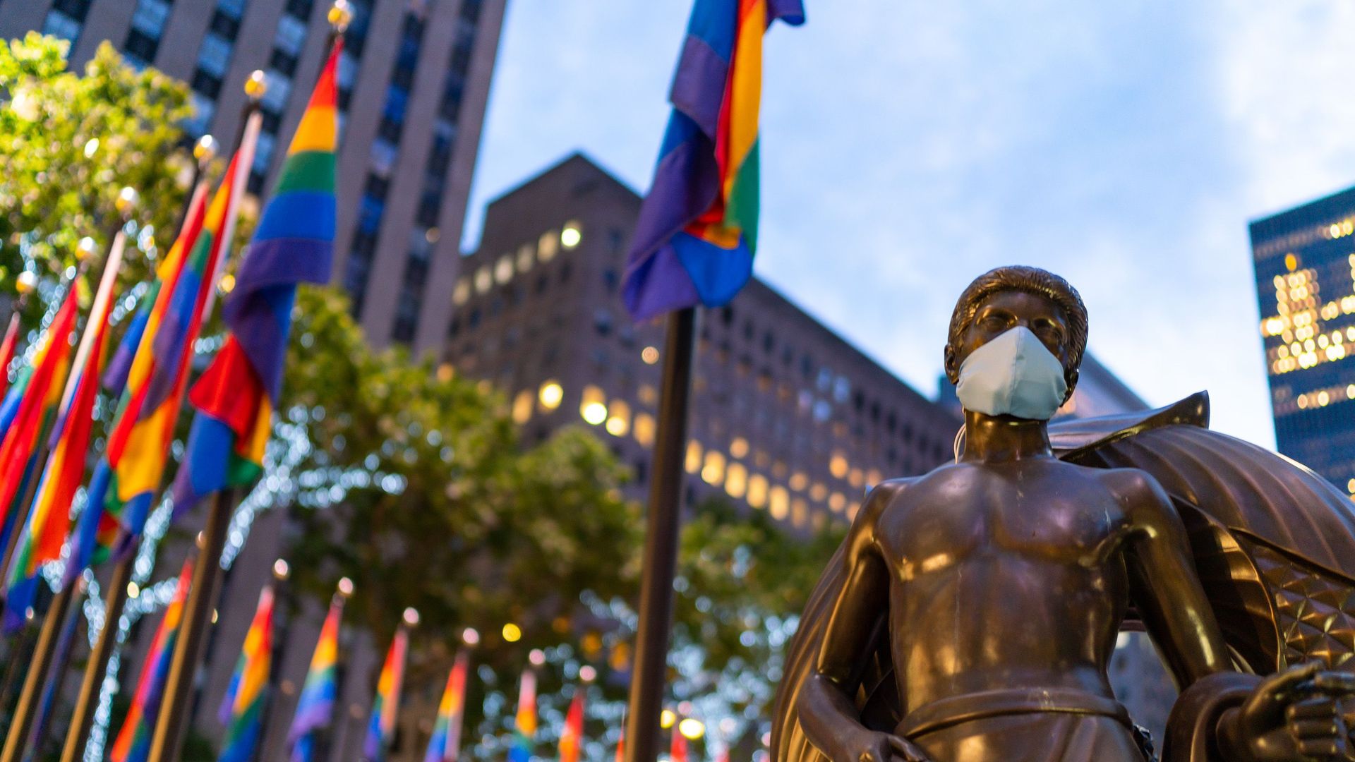 A statue wears a face mask with rainbow flags behind it 