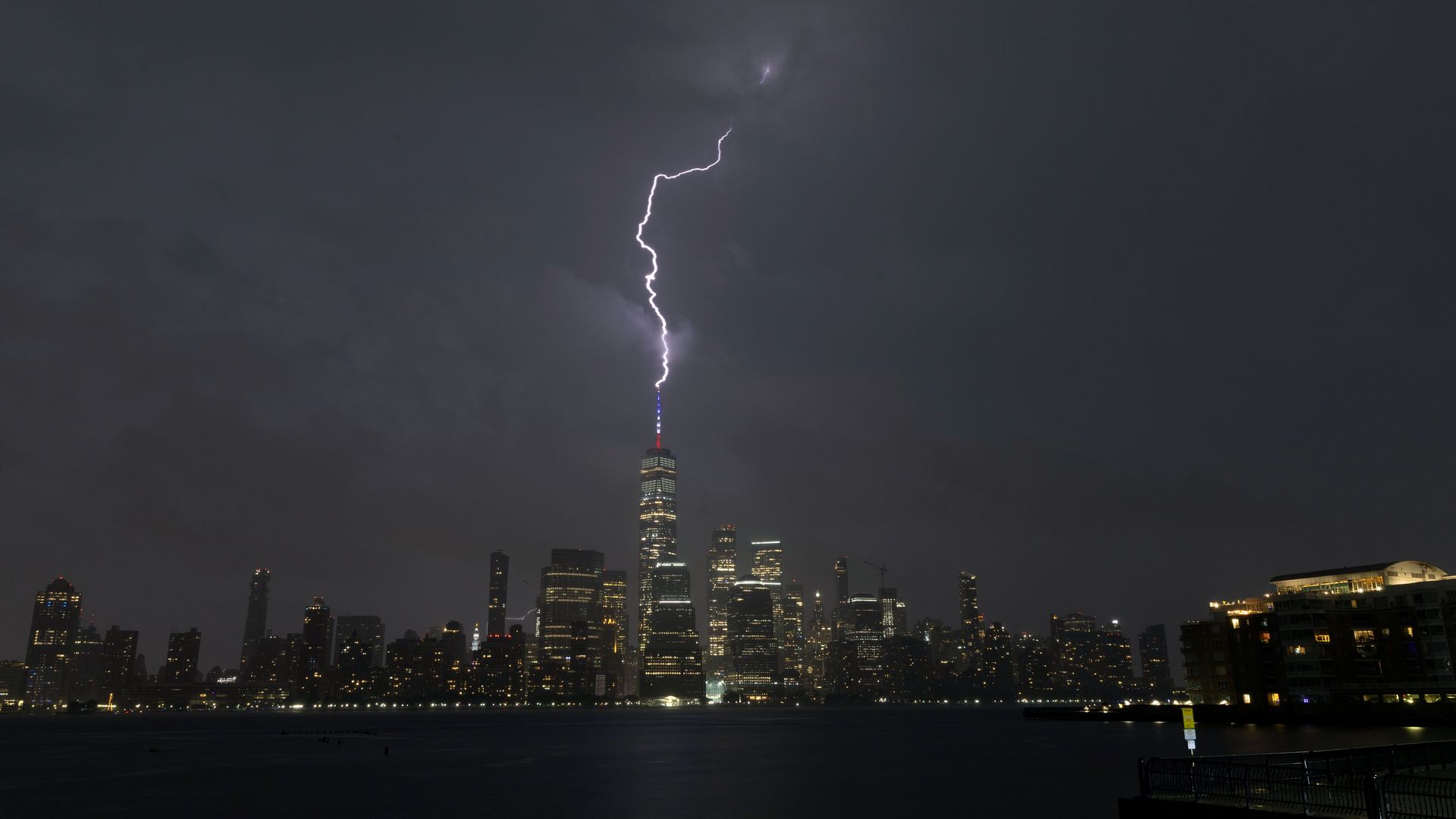 Nighttime city skyline along the water as a bright lightning bolt descends from dark clouds and strikes the tallest central skyscraper, whose antenna is lit red and blue.