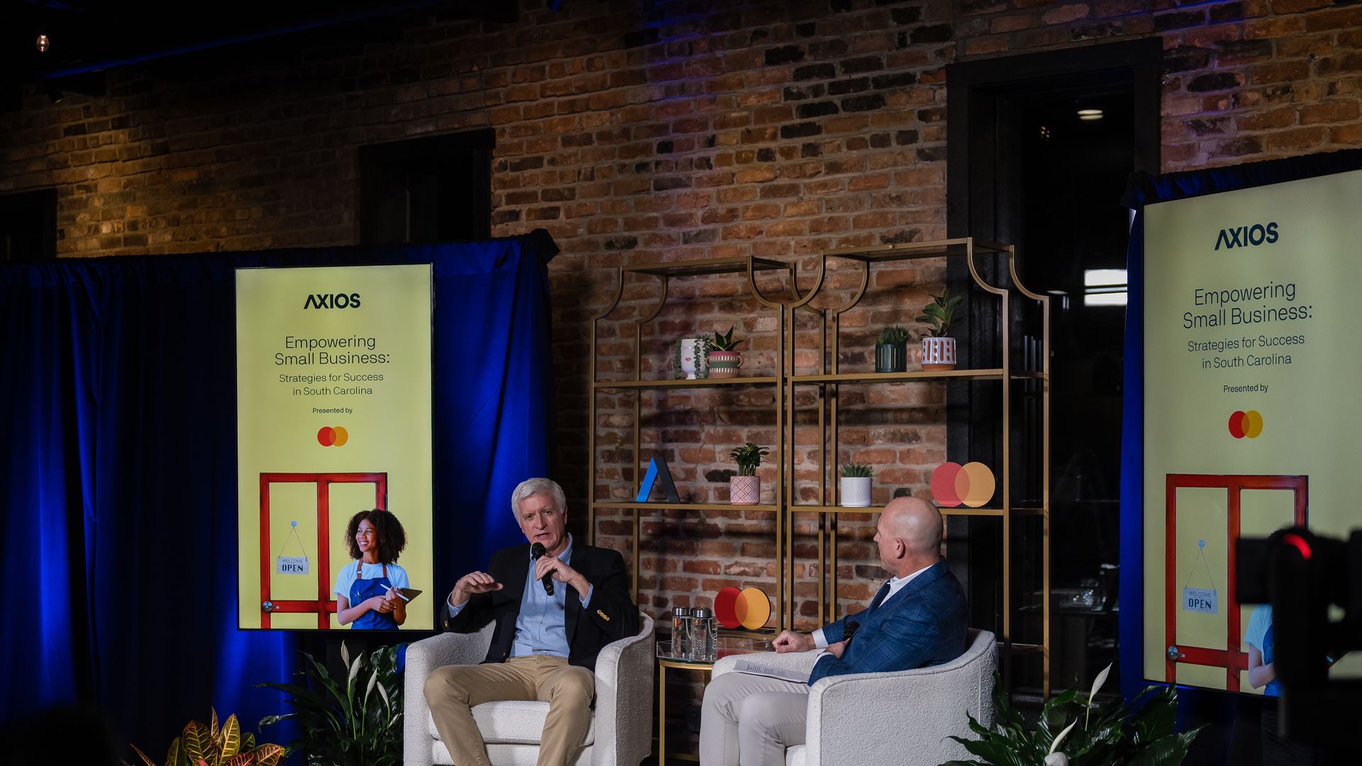 Two men seated on stage holding microphones during an Axios event titled "Empowering Small Business: Strategies for Success in South Carolina," with brick wall, plants, and display screens in background.
