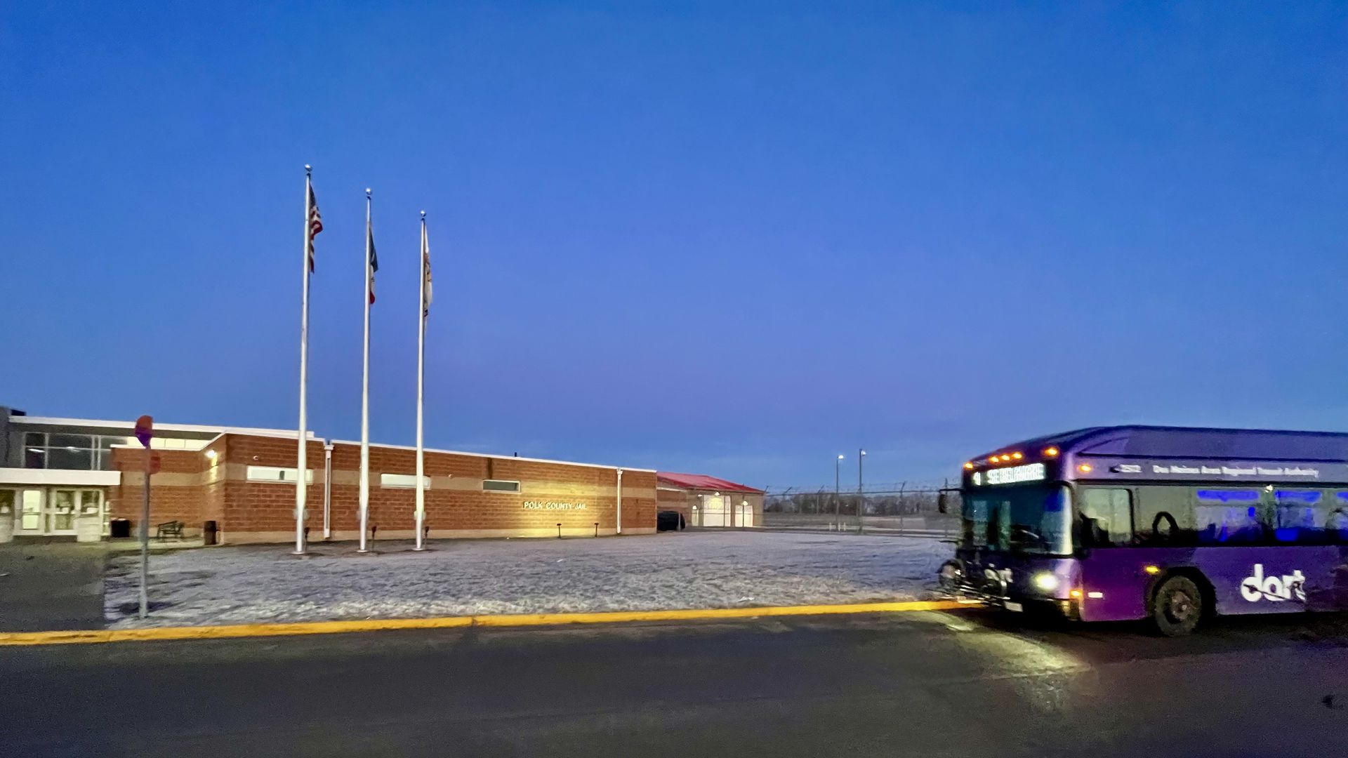 Purple Dart transit bus with lights on parked near brick Polk County Jail building at dusk, three flagpoles with flags, and a chain-link fence in background.