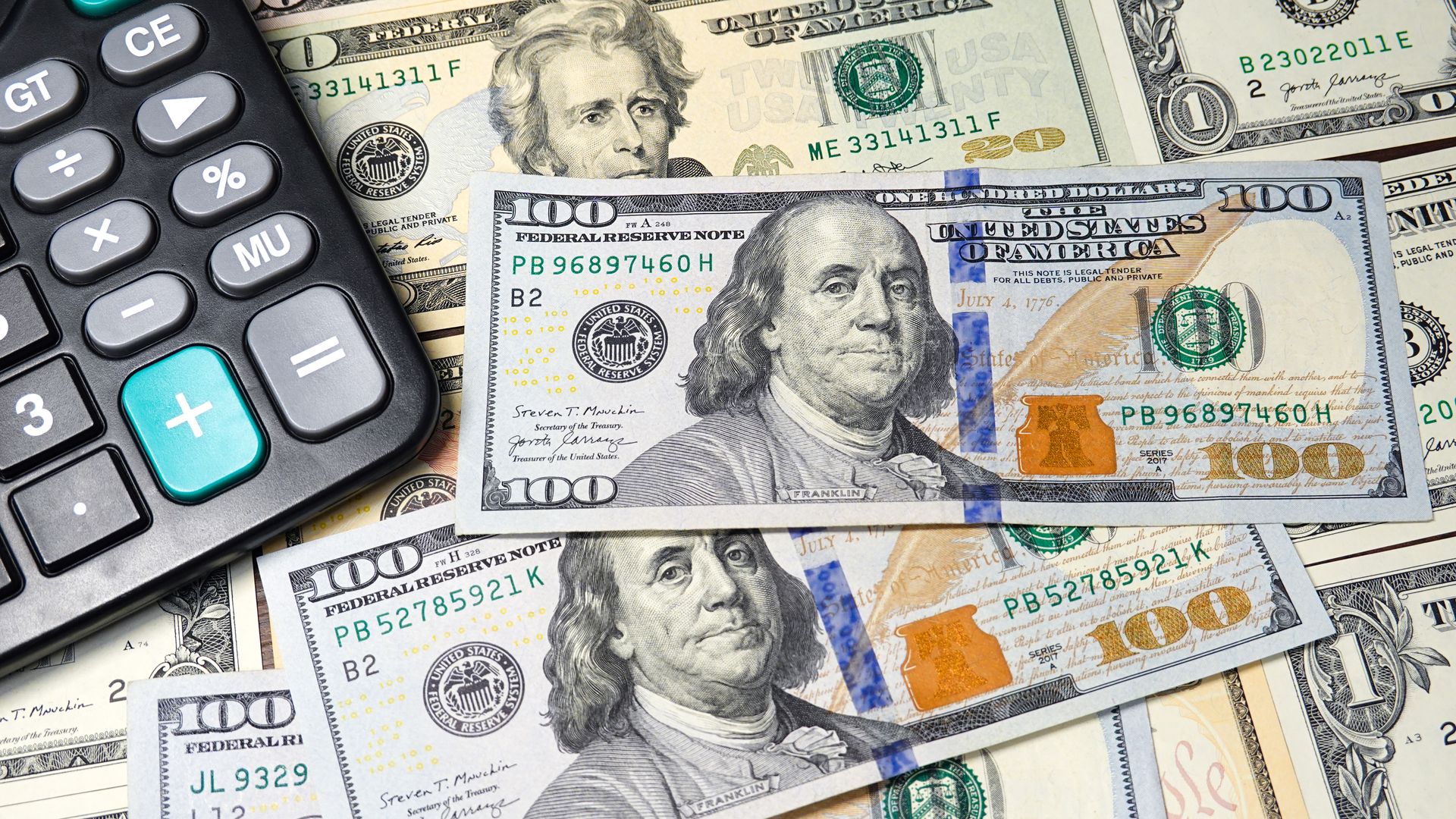 U.S. currency, including $100 bills, is strewn across a surface with a black standard calculator visible in the top left corner. 