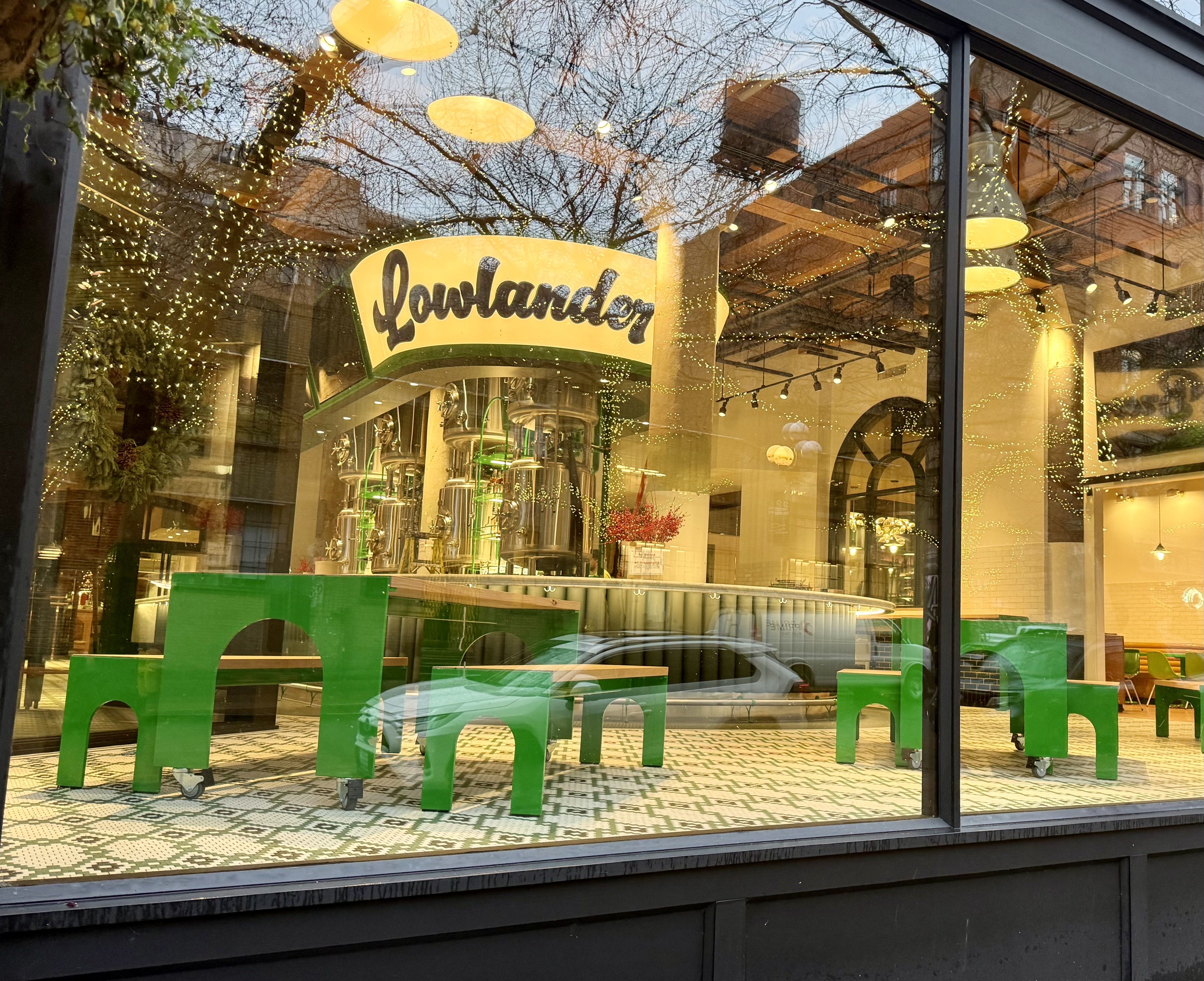 Reflection of street and trees in a window showing green tables and stools inside a warmly lit brewery named Lowlander with decorative lights and tiled floor.