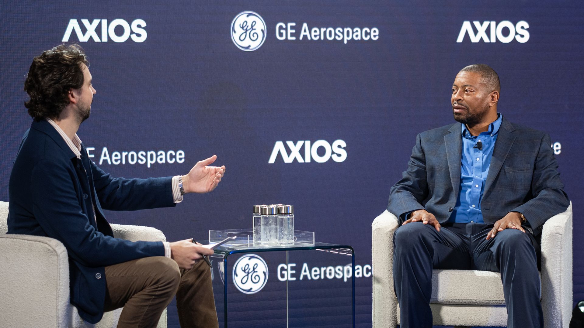 Two men in suits sit in cream chairs on a stage with a blue backdrop featuring AXIOS and GE Aerospace logos; one gestures while the other listens, a glass coffee table with water bottles between them.