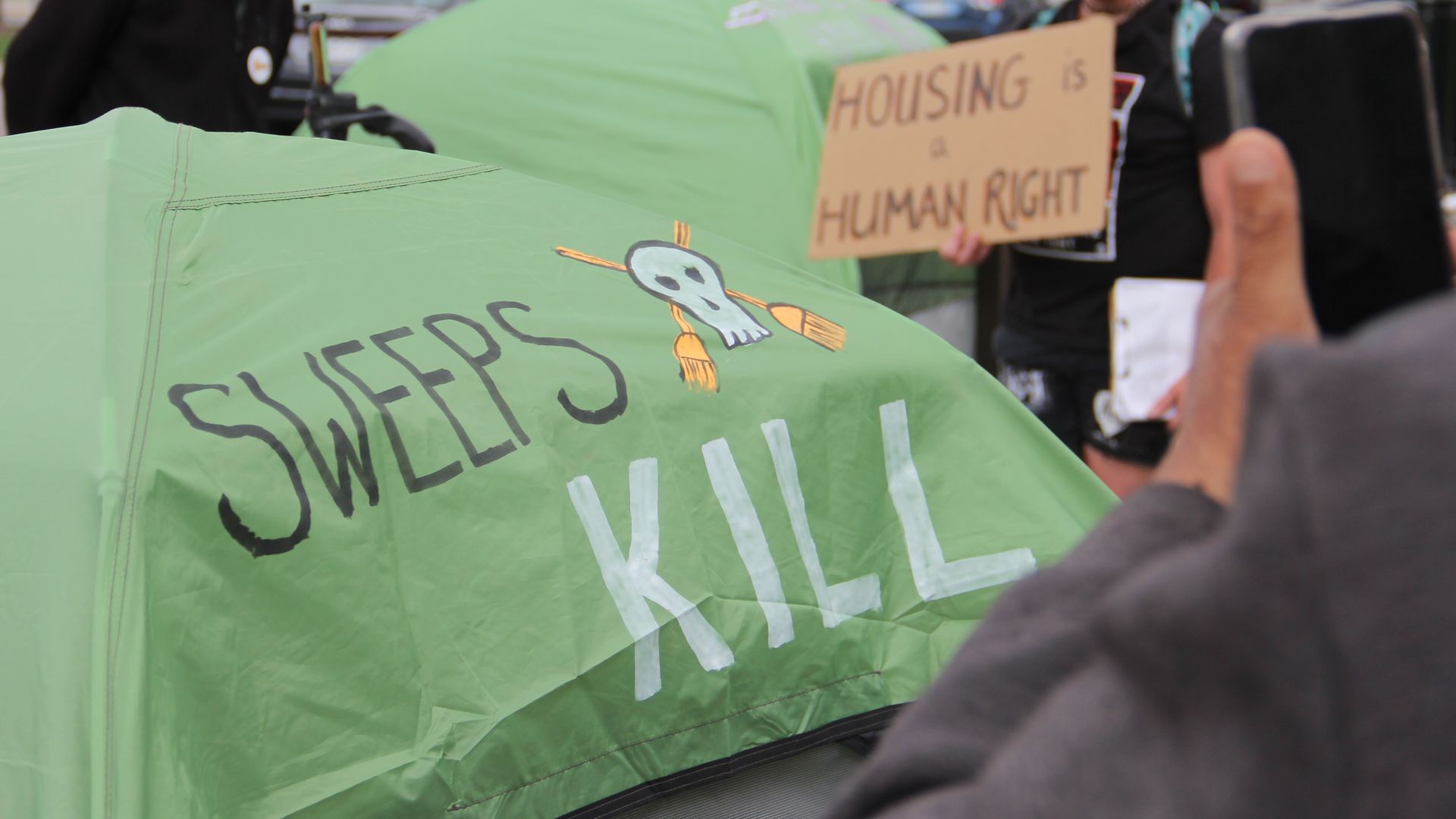 A green camping tent with the words SWEEPS KILL written on it, with a small skull cartoon next to it. In the foreground, a person is taking a photo with their phone.