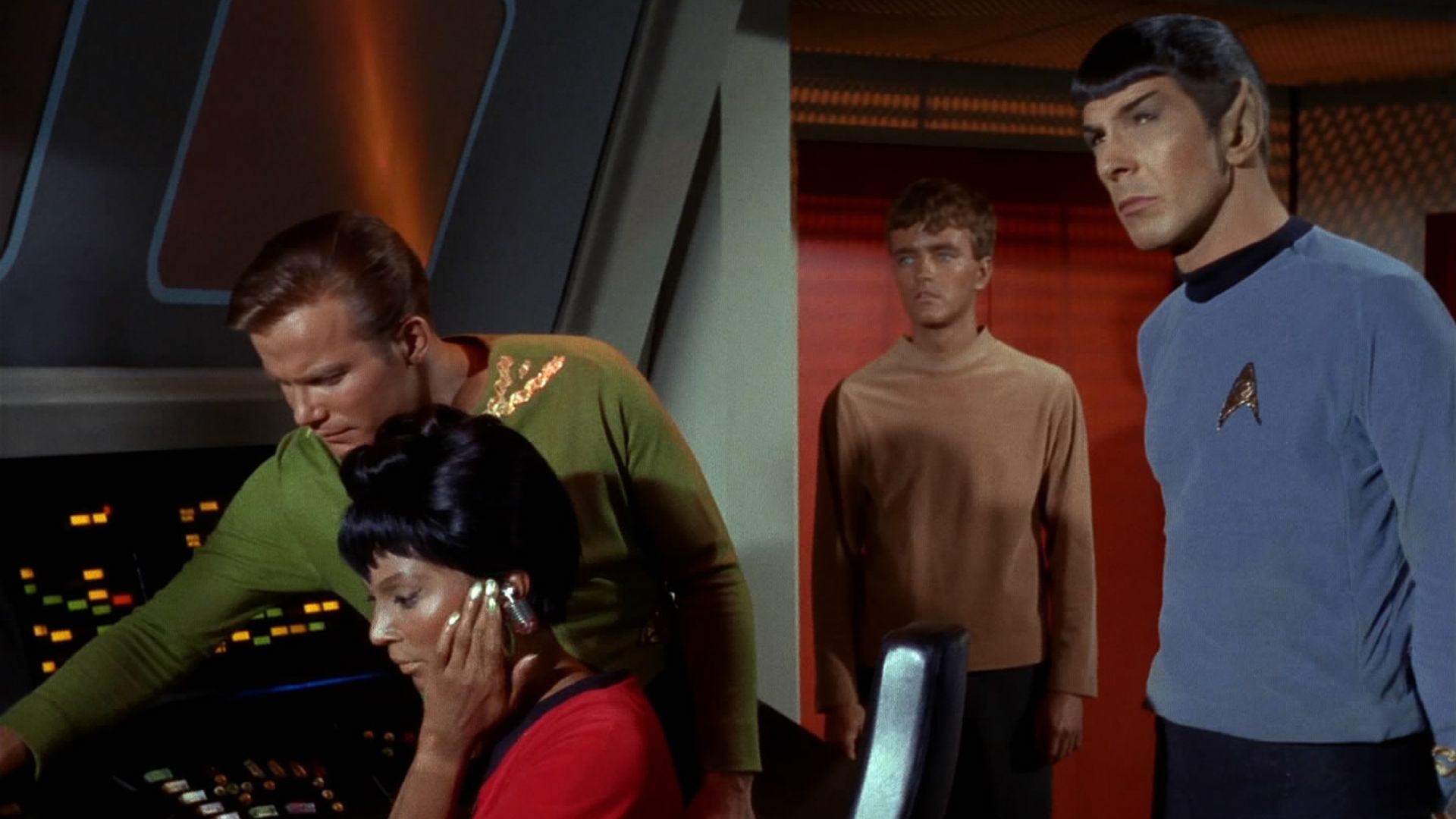 Spaceship officers in green, red, beige and blue uniforms stand at a command center