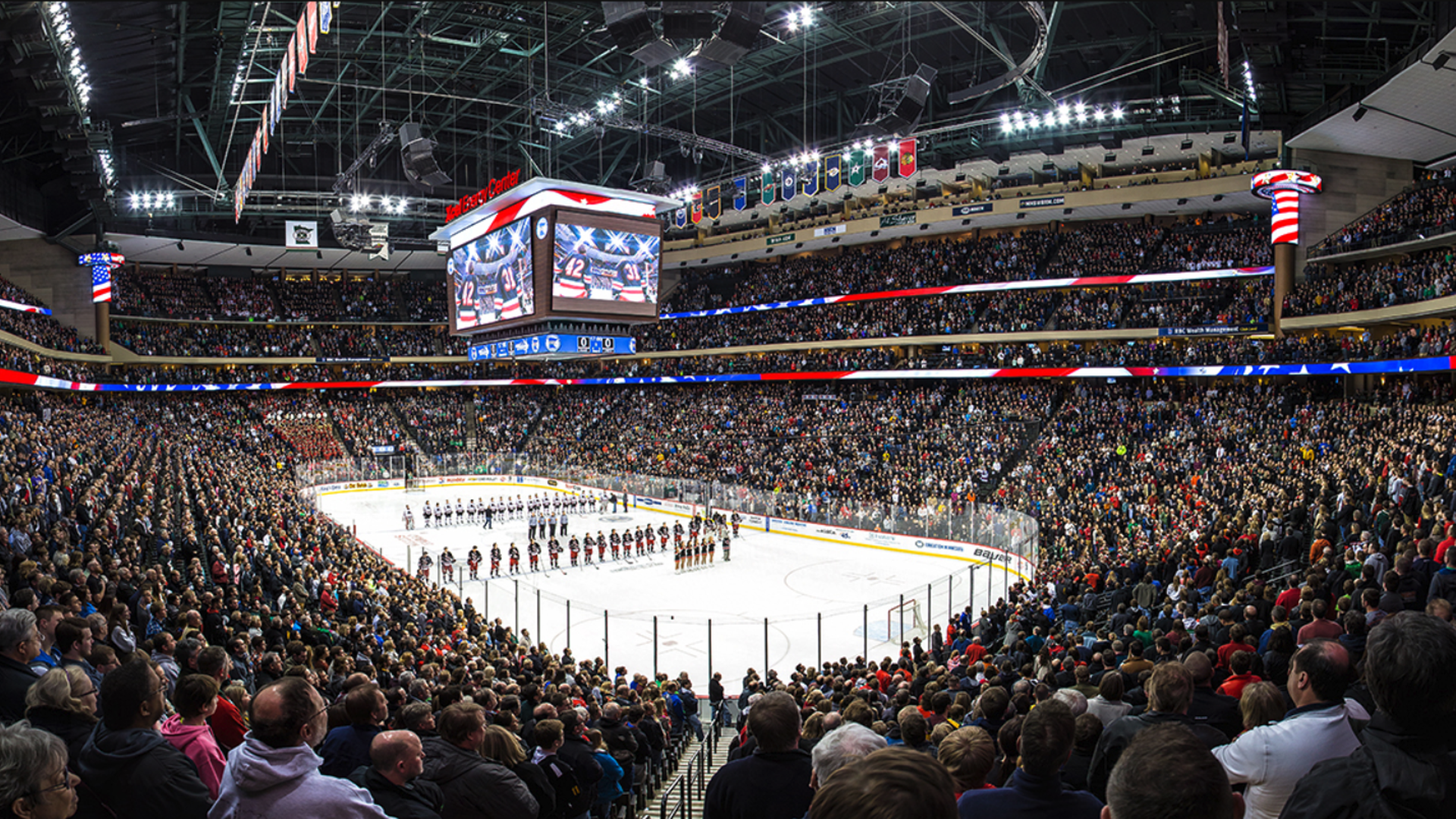 A professional hockey arena full of spectators stands during the national anthem at a high school hcokey game.