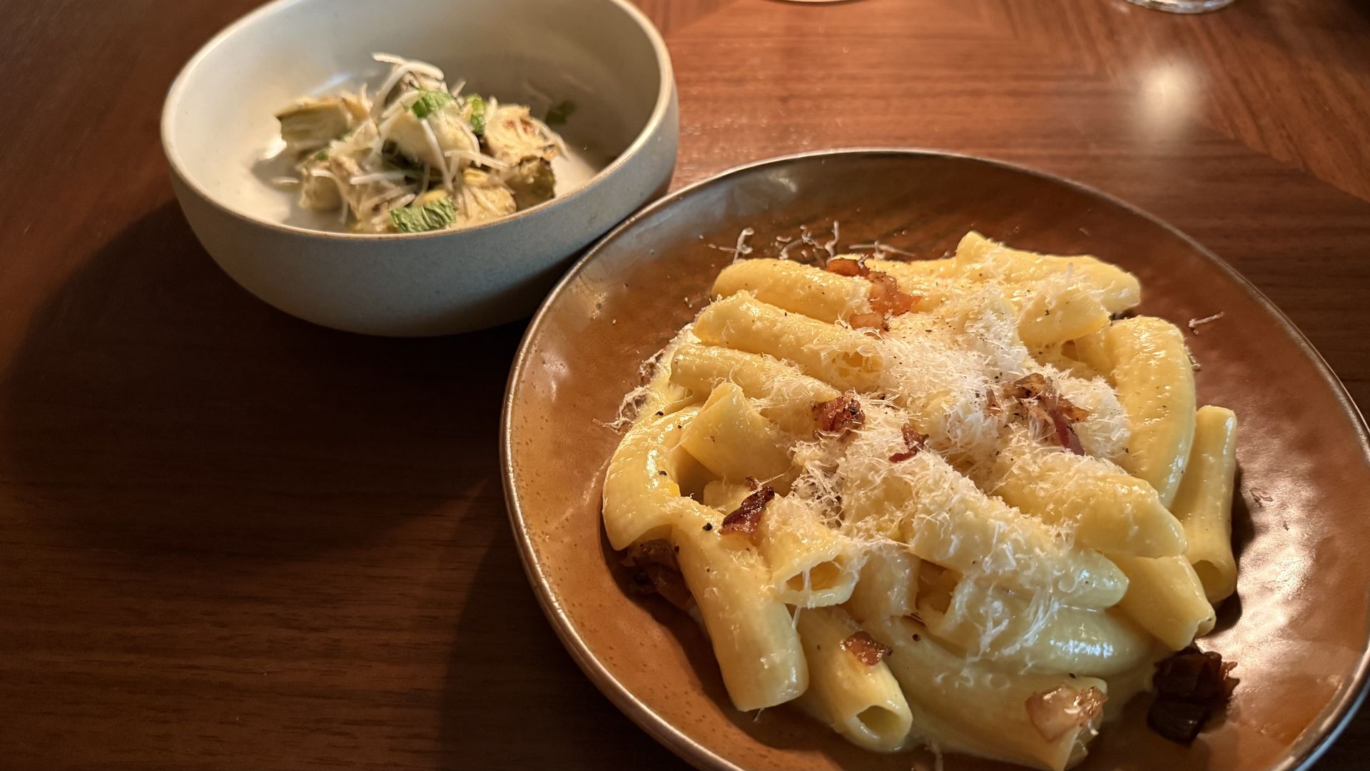 Two dishes on a wooden table: a brown plate of creamy pasta with grated cheese and crispy bits of meat, and a white bowl with shredded cheese and herbs over grilled artichoke hearts.