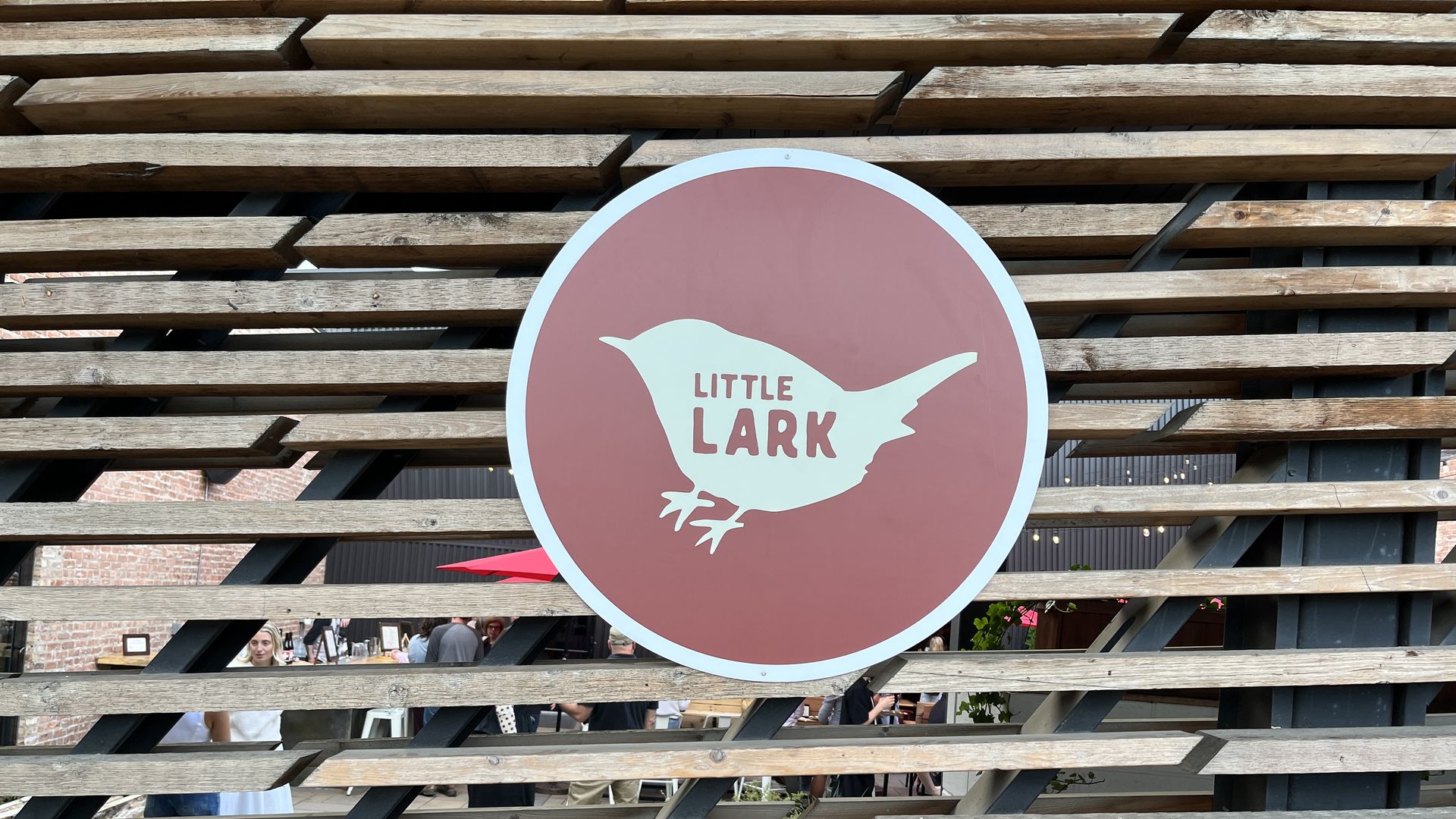 Round pink sign with a white bird silhouette and the words "Little Lark" mounted on a slatted wooden fence, with people and outdoor umbrellas visible behind it.