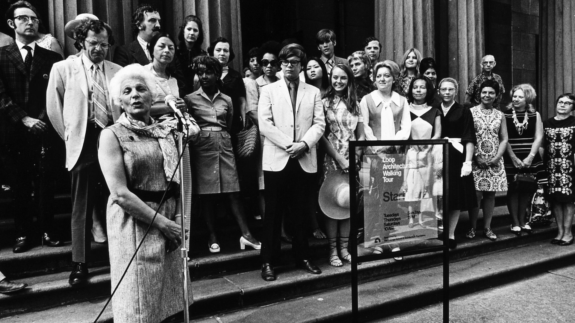 Black-and-white group photo on building steps; an elderly woman speaks into a microphone at a stand, while a framed poster advertises a walking tour beside the crowd.