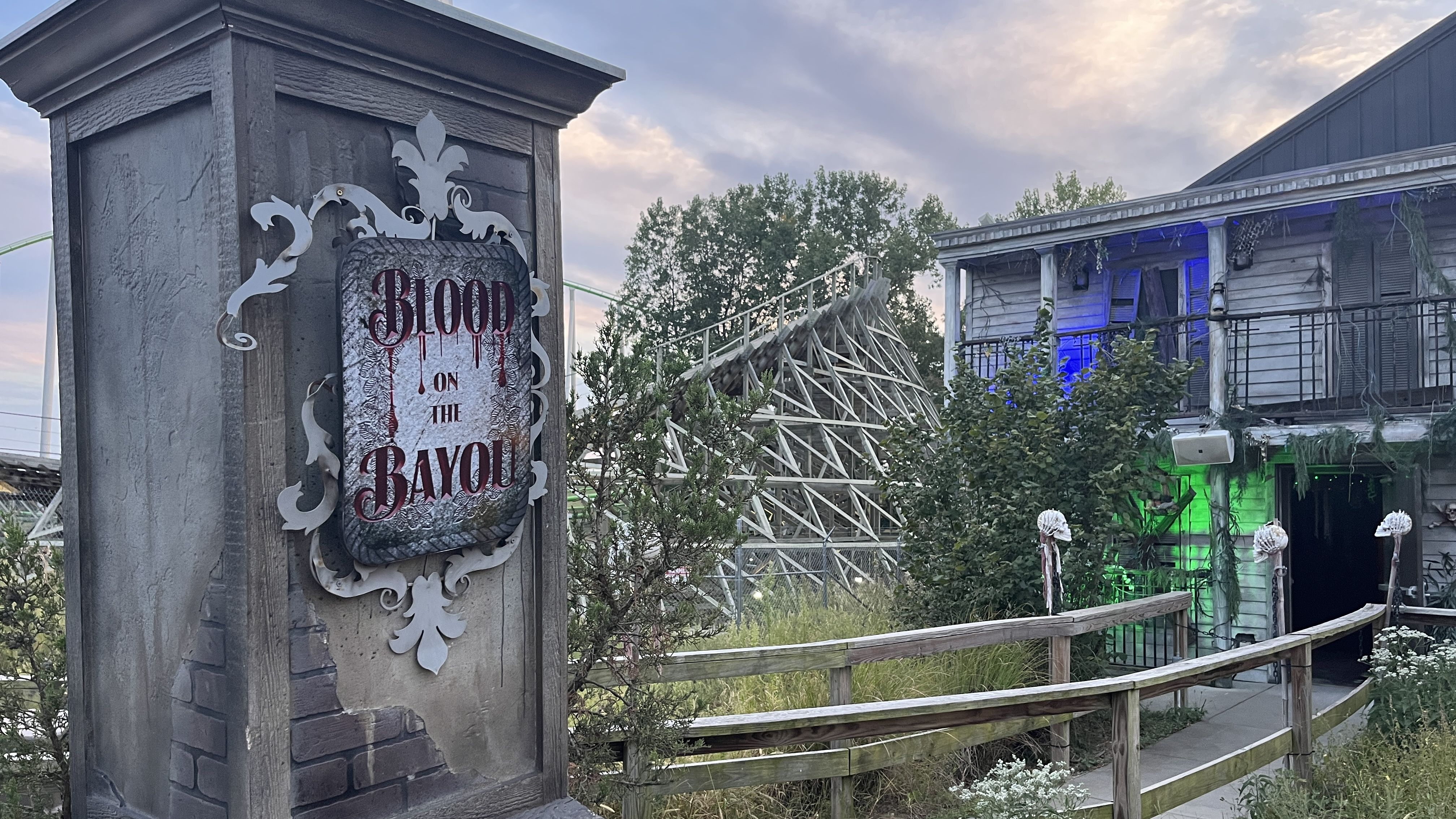 Sign reading "Blood on the Bayou" on a weathered stone pillar with gothic decorations; behind is a wooden roller coaster and a spooky two-story house with green and blue lights amid trees and plants.