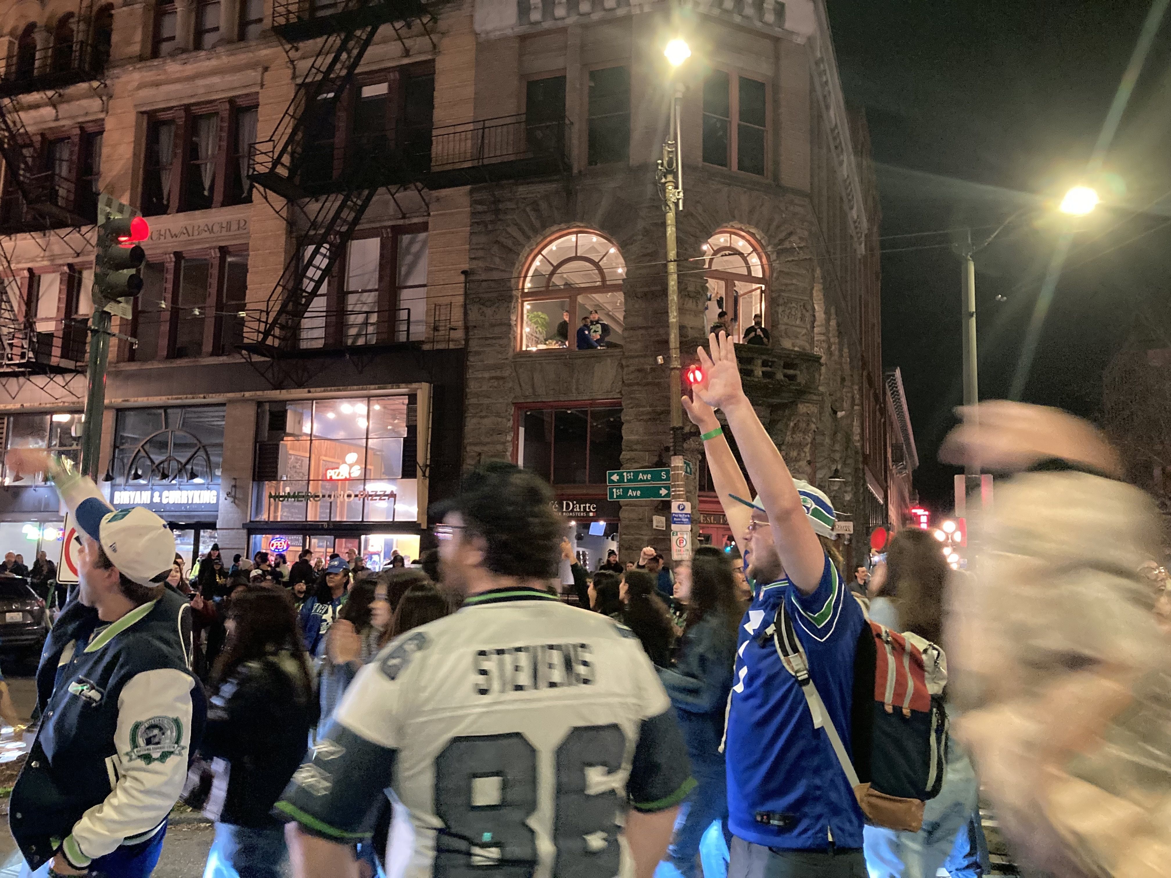 Nighttime street scene with a crowd, some wearing blue and white Seahawks jerseys, celebrating near old brick buildings with people watching from upstairs windows.
