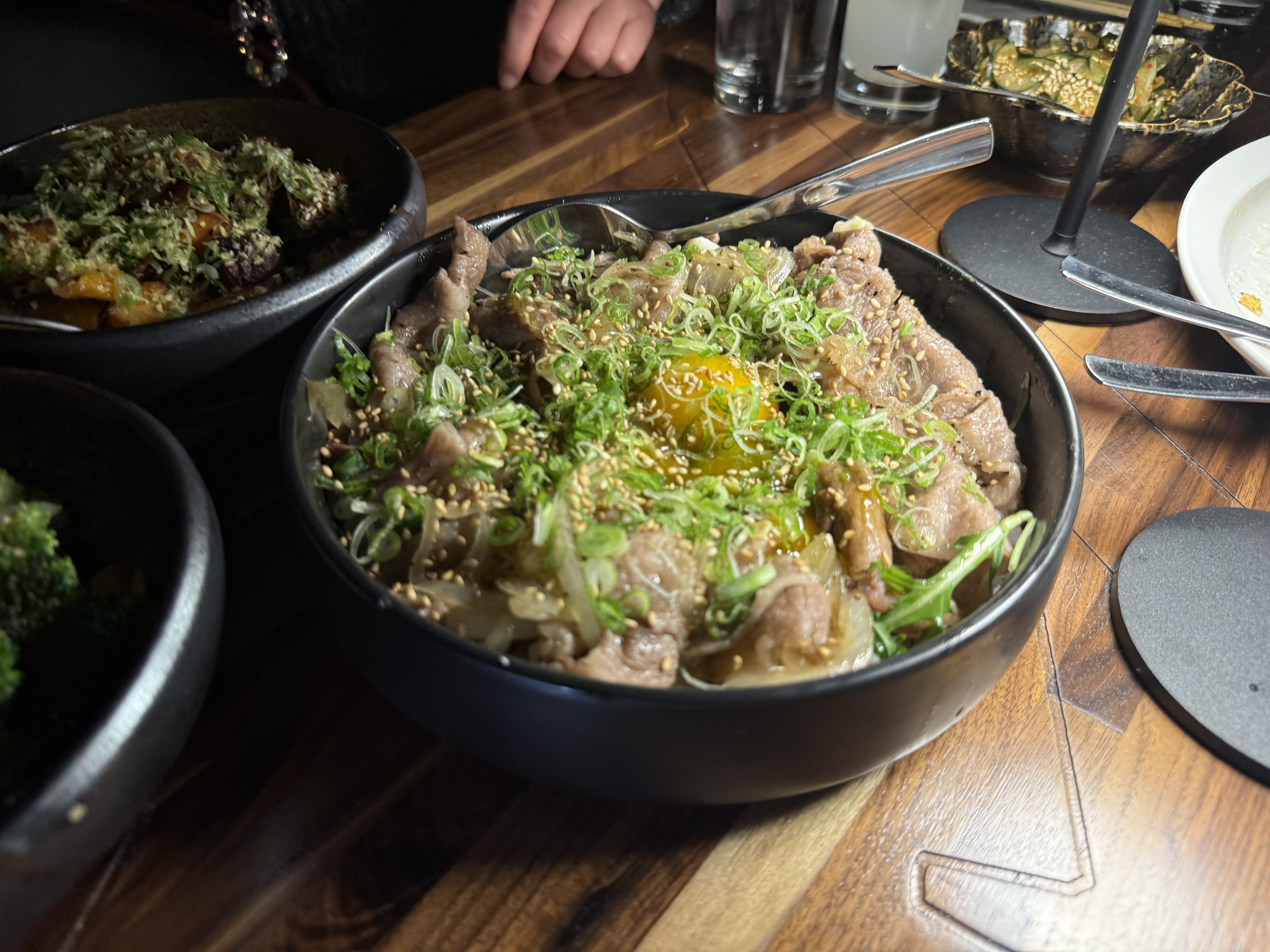 Black bowl on wooden table with beef slices topped with green onions, sesame seeds, and a raw egg yolk in the center. Other black bowls with food and a white plate nearby.
