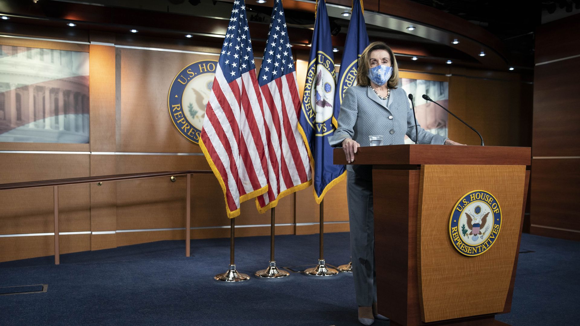 Photo of a masked Nancy Pelosi standing at a podium with the American flag behind her