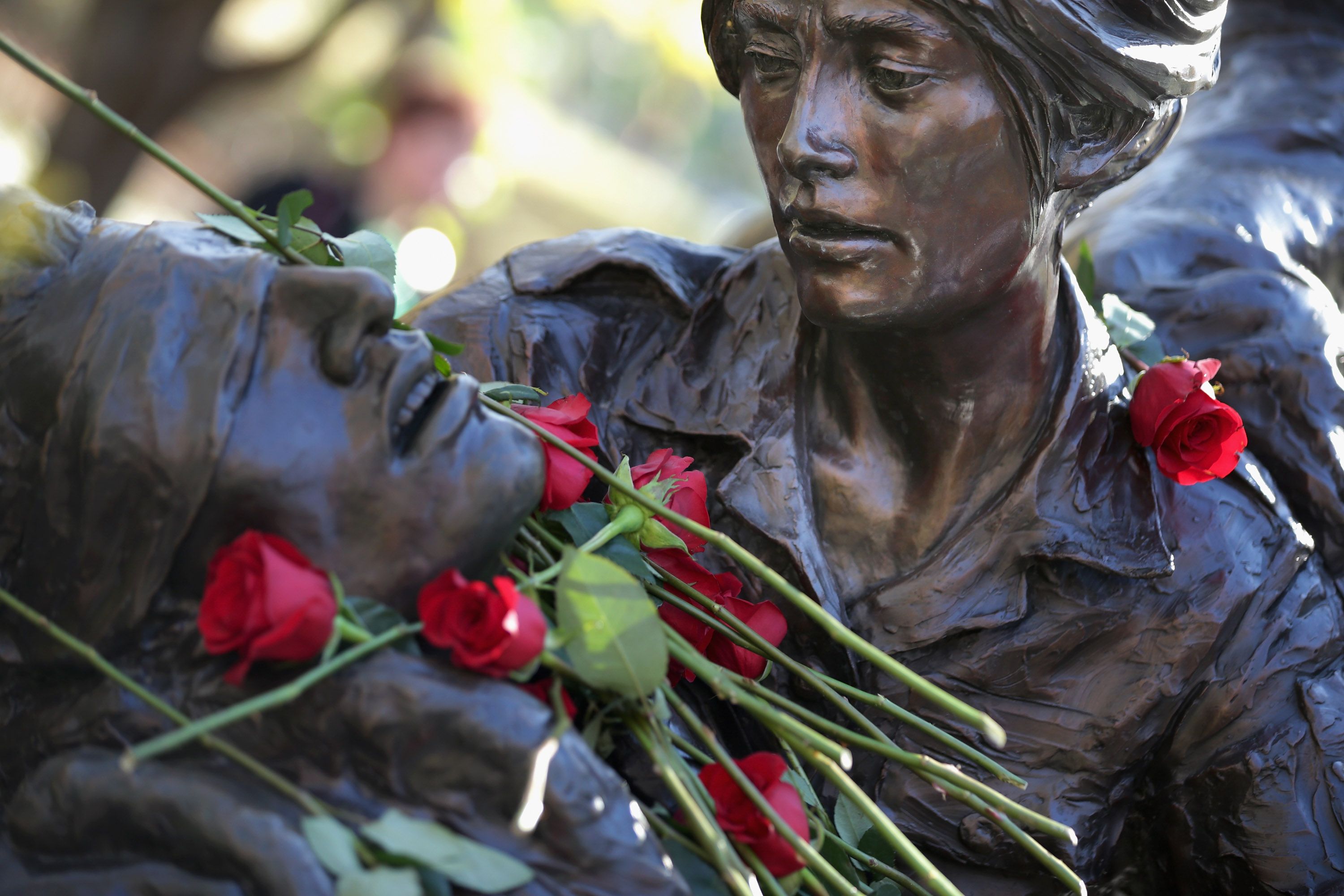 Close-up of a bronze statue with arms outstretched, dark patina, red roses laid across its chest and arms, with a blurred green background.