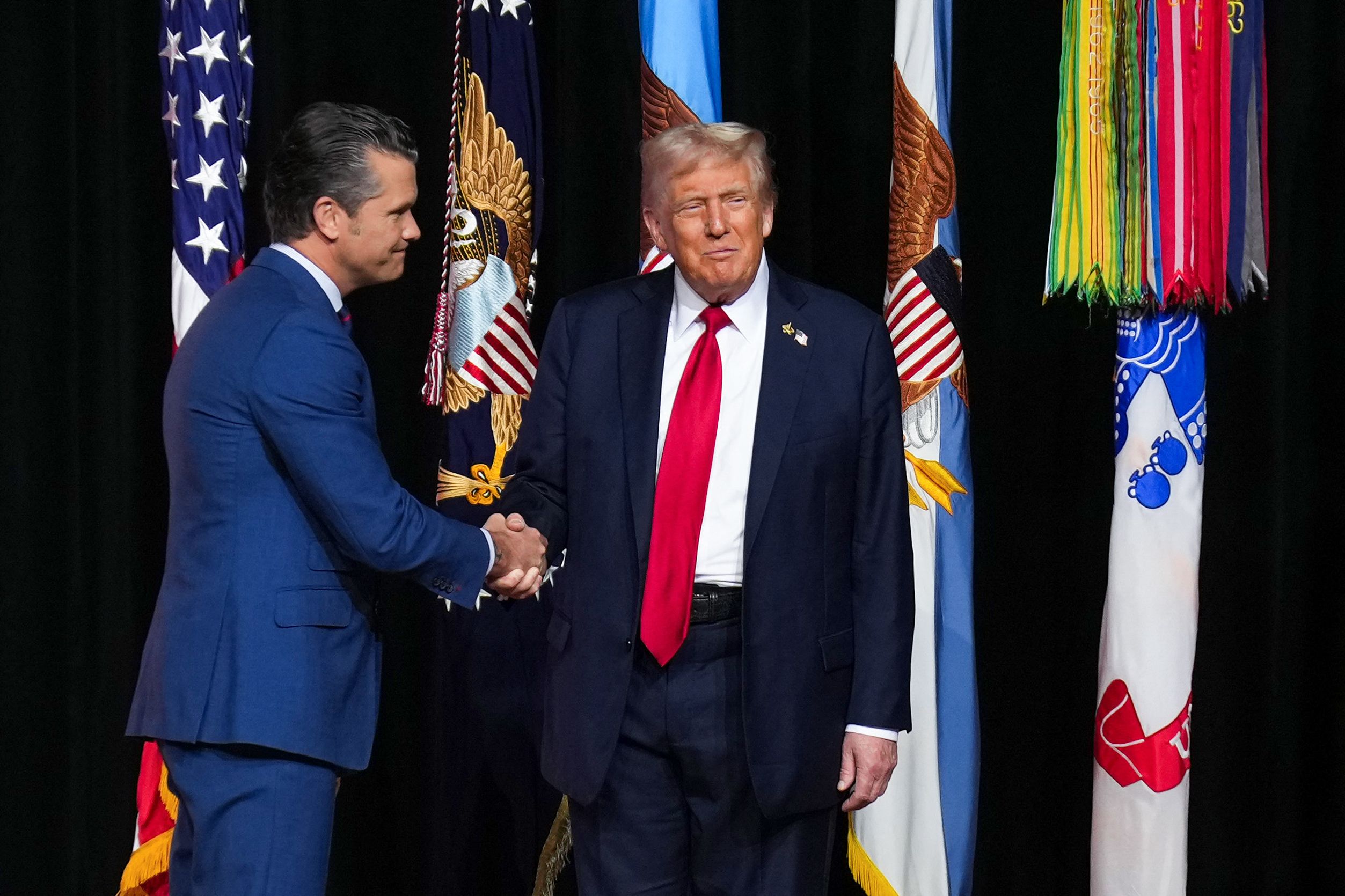 Two men shake hands in front of flags on a stage. 