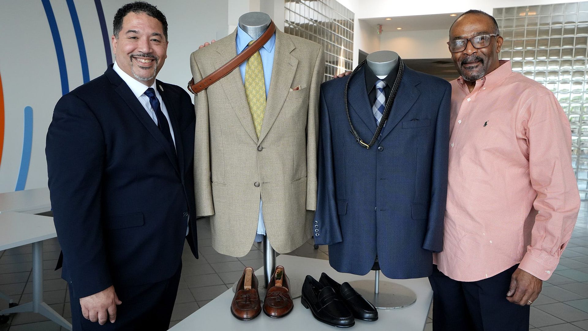 Two men stand beside suit displays at a nonprofit offering low-income men with professional warddrobes.
