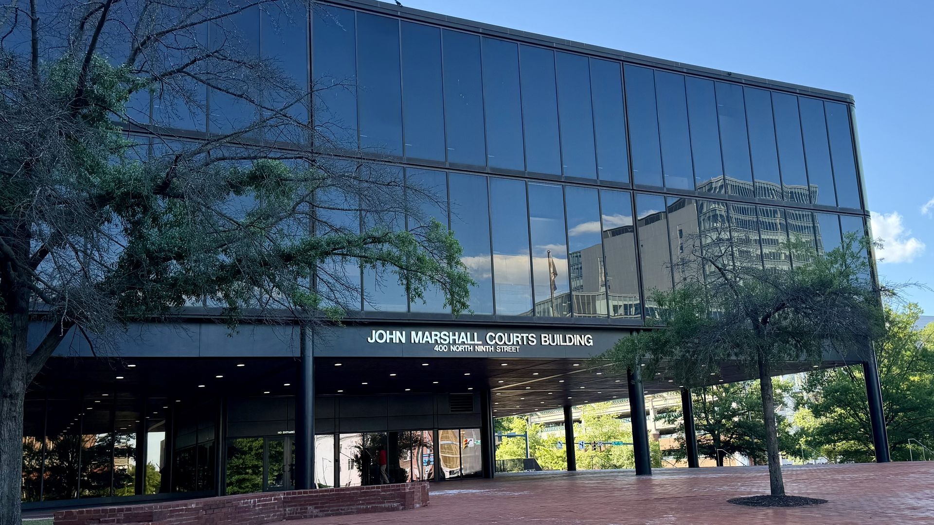 Front view of John Marshall Courts Building, a glass blue-tinted facade reflecting the sky and trees, with a sign reading "JOHN MARSHALL COURTS BUILDING, 400 NORTH NINTH STREET".
