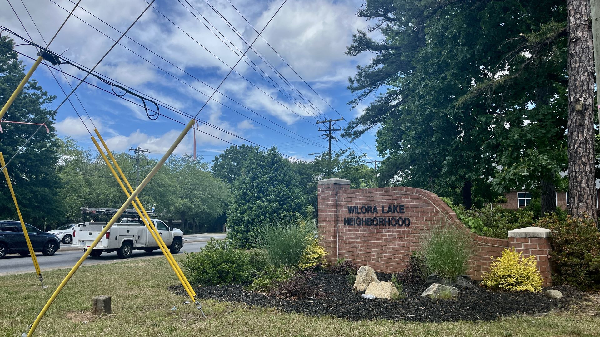 Wide street scene with crisscrossing overhead power lines and leaning yellow utility poles. A brick sign reads "WILORA LAKE NEIGHBORHOOD" beside green shrubs and trees, with parked cars under a blue, cloudy sky.