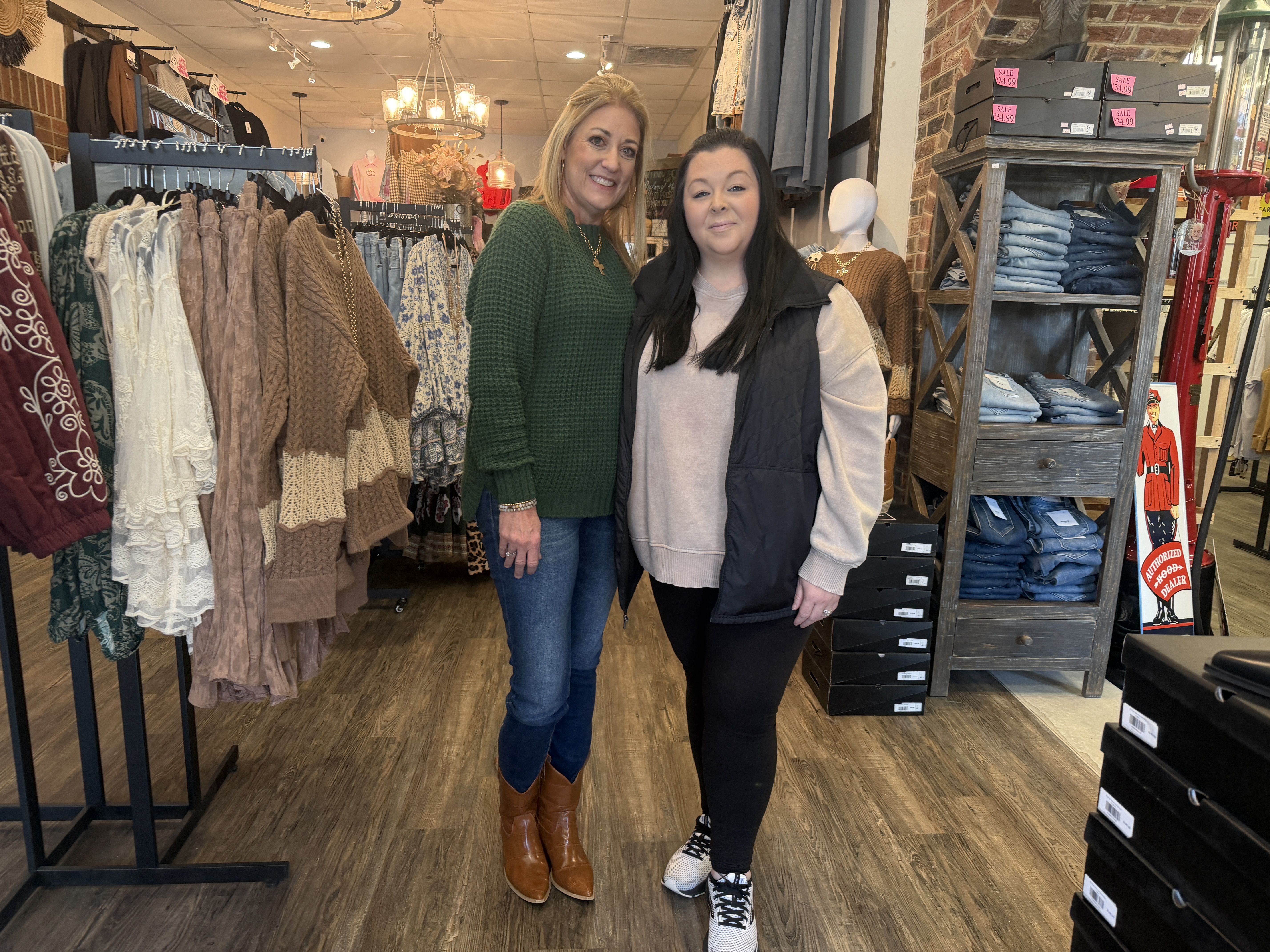 Two women standing inside a clothing store; one wears a green sweater and brown boots, the other in a beige sweatshirt, black vest, and sneakers; racks of sweaters and jeans around them.