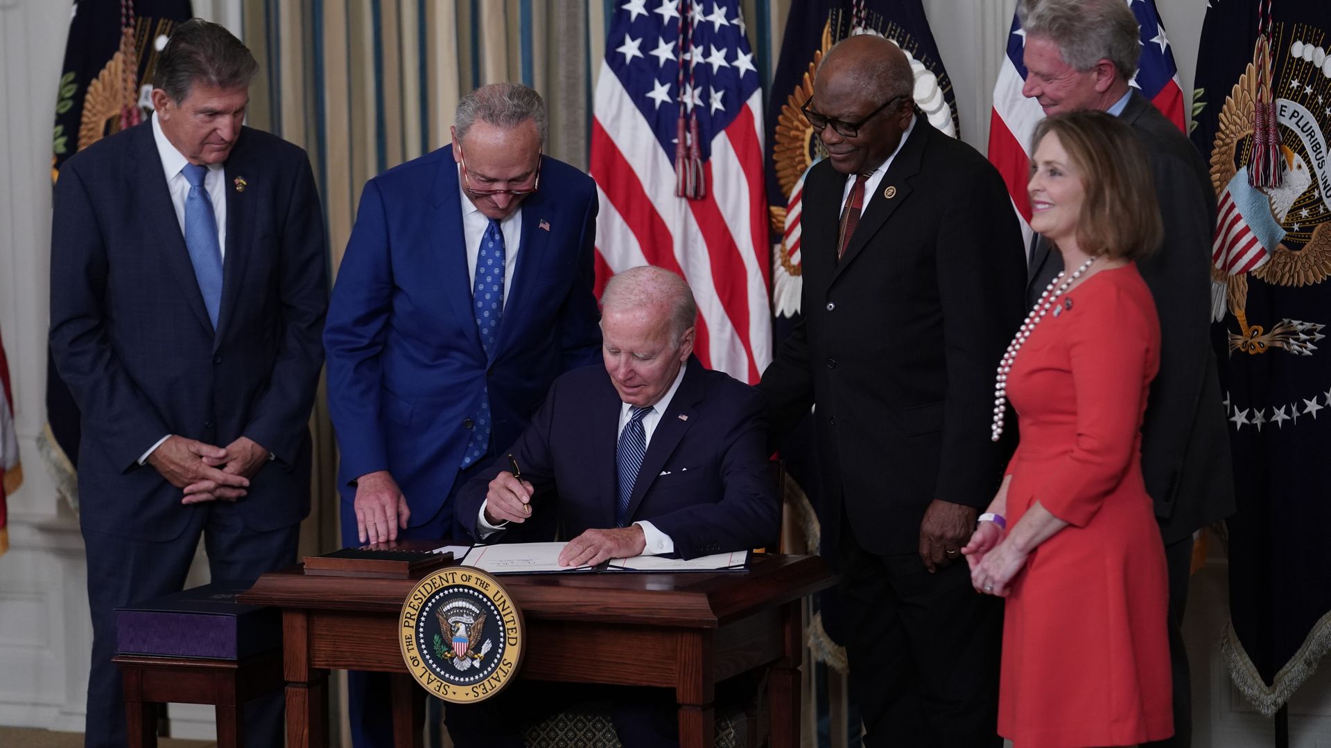 Photo of Joe Biden signing a bill on a desk with lawmakers crowded around and behind him