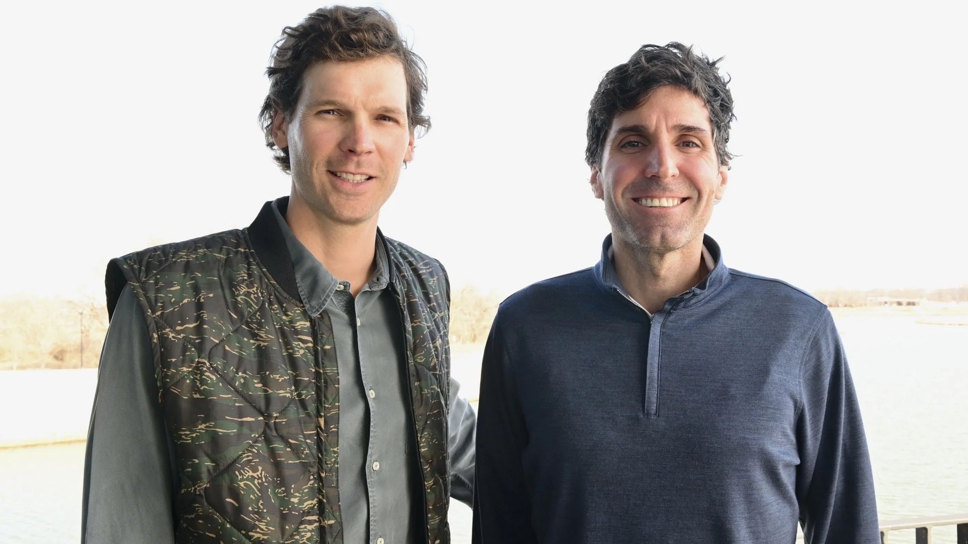 A photo of two men standing side-by-side on a balcony with the outdoors visible in the background.