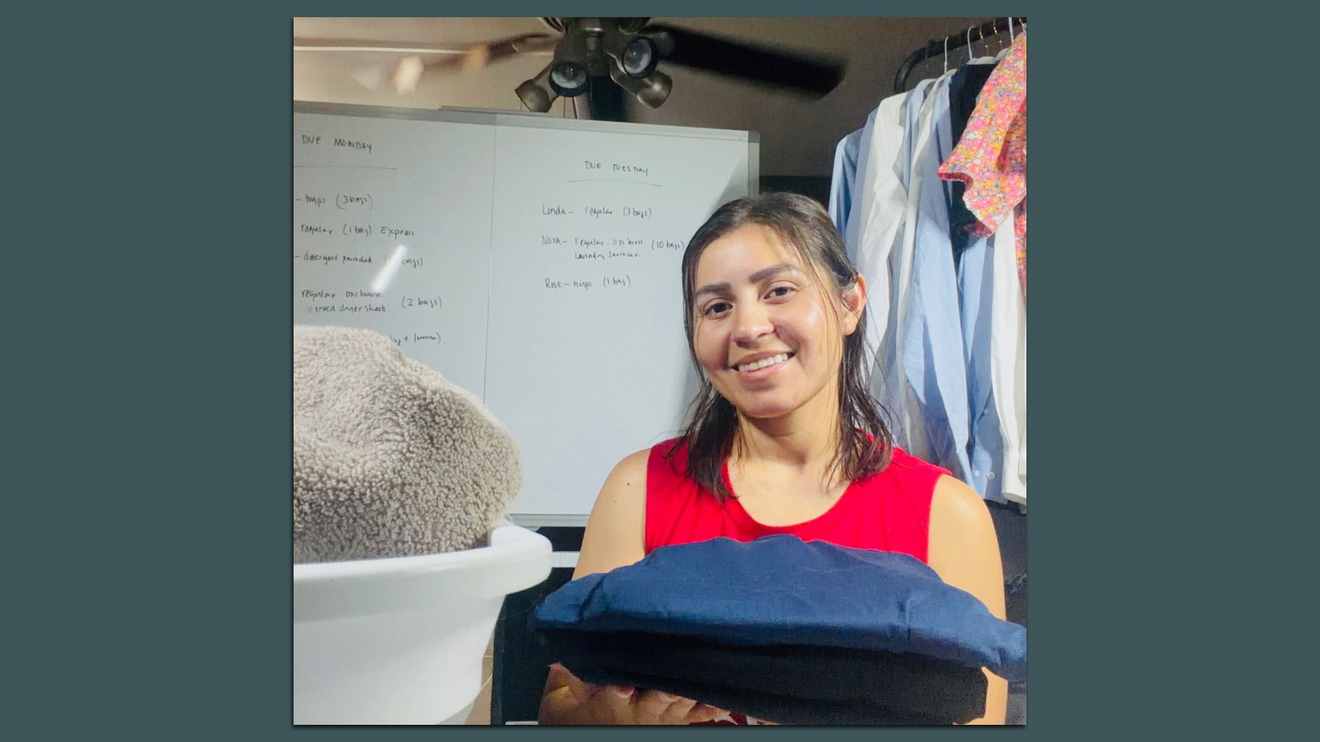 Smiling woman in a red sleeveless top sits in a cluttered room with a whiteboard behind her, clothes on a rack to the right, and a ceiling fan overhead; a beige fuzzy item sits in the foreground.
