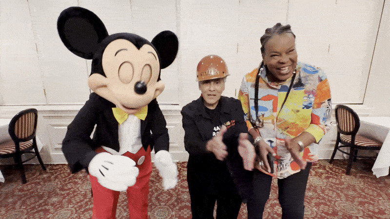 Photo shows Mickey Mouse dancing with Stella Chase Reese and Disney Imagineering exec Charita Carter.