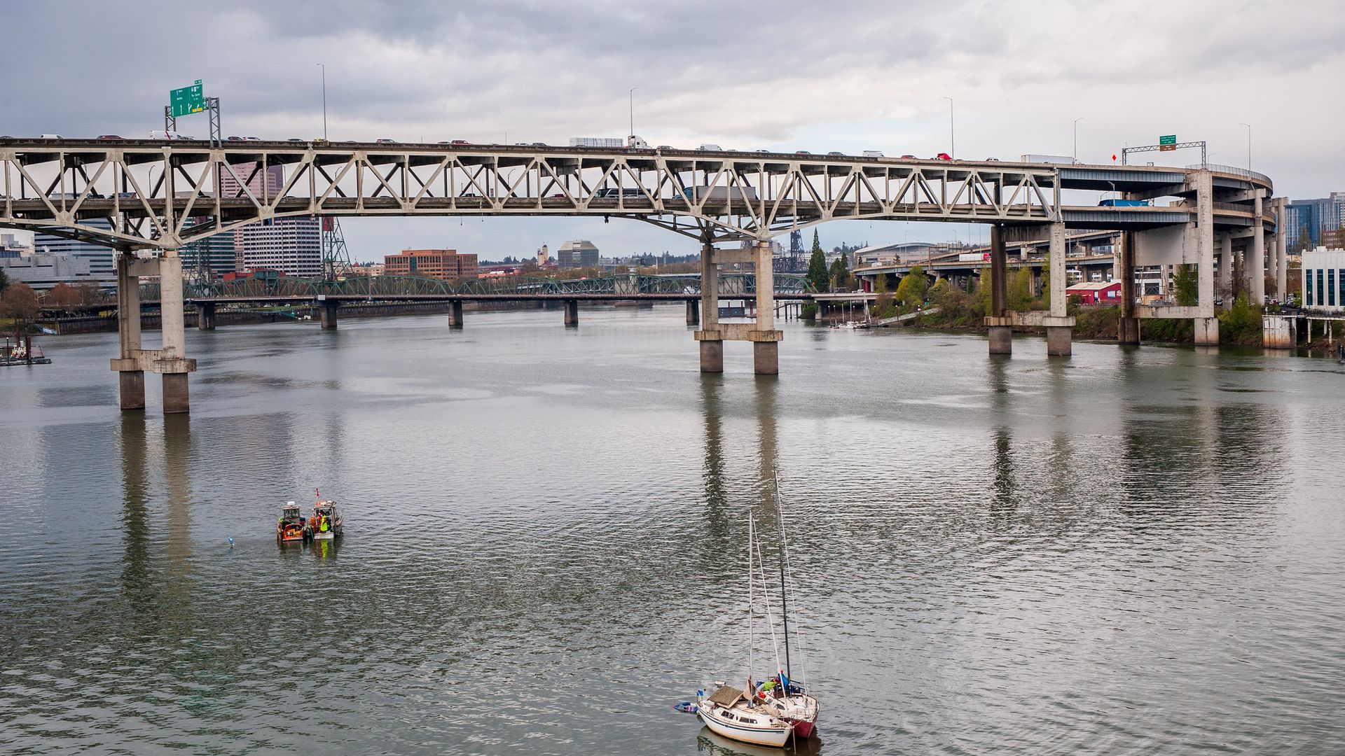 Gray steel bridge with multiple vehicles crosses a river with two small boats in the water, city buildings and cloudy sky in the background.