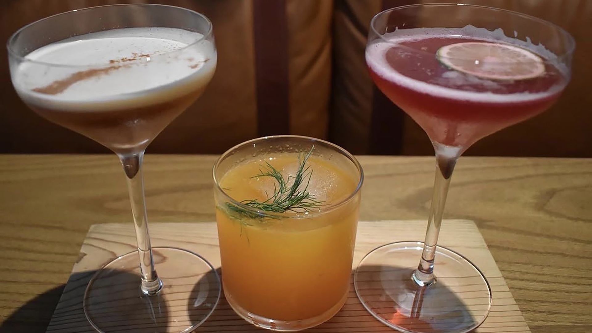 Three cocktails on a wooden surface: left a creamy white drink with cinnamon dusting in a coupe, center an orange drink with dill garnish, right a pink-red cocktail with a lemon slice and sugared rim.