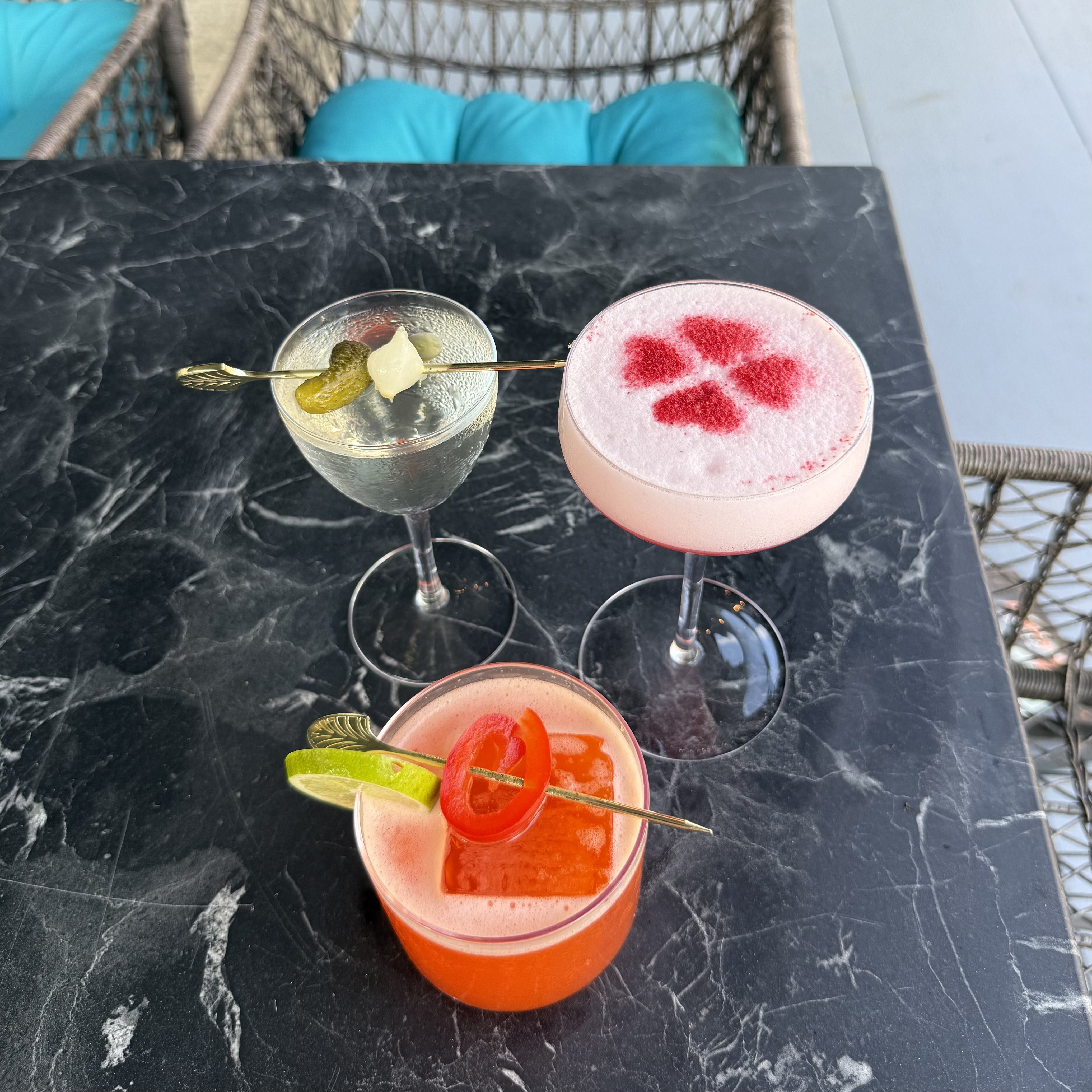 Overhead view of three cocktails on a black marble table: a clear drink with a pickle skewer, a pink foamy drink with red powder, and a red drink with lime; teal cushions in the background.
